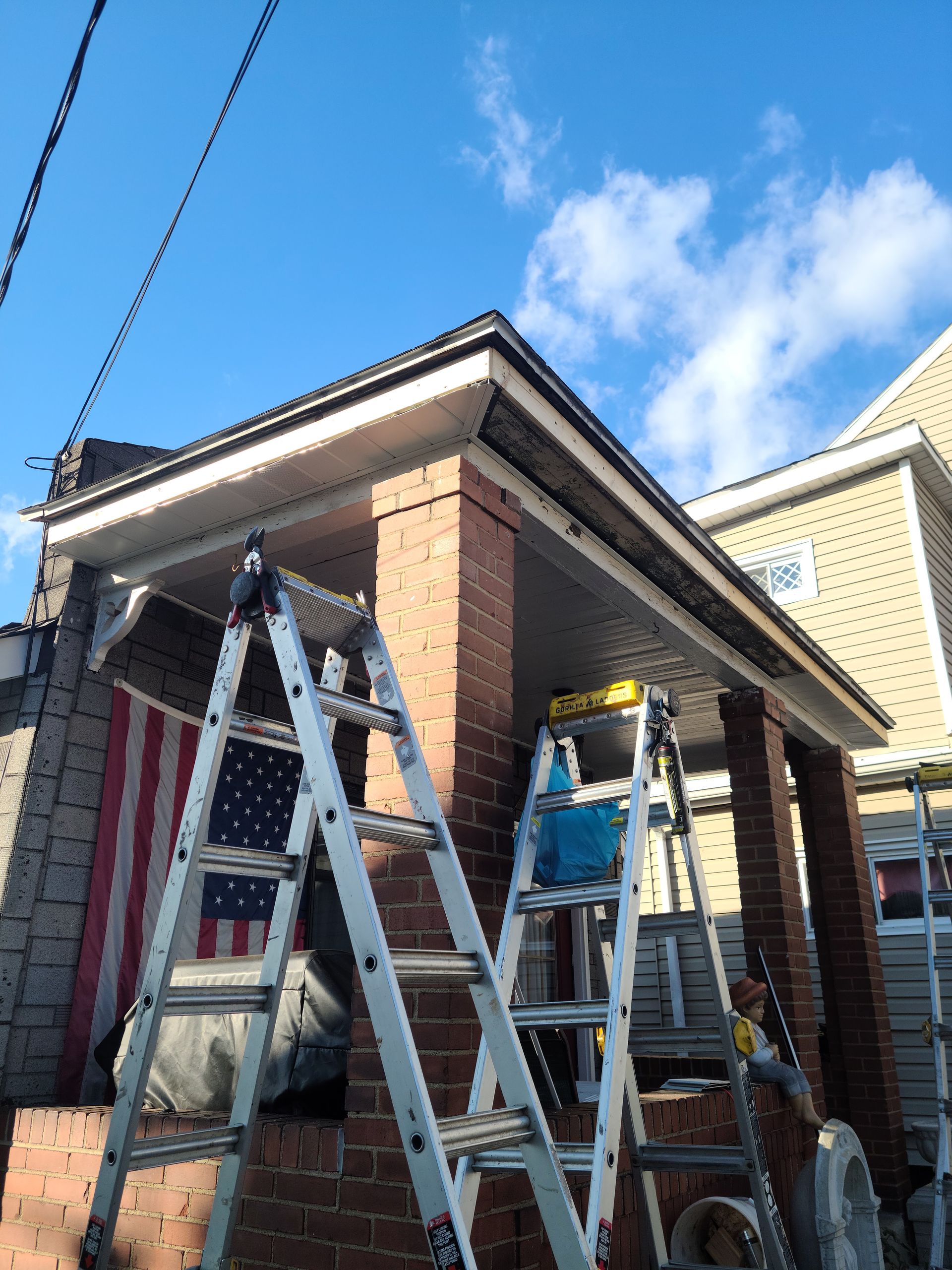Two ladders propped against a brick porch with a partially visible American flag; bright sky.