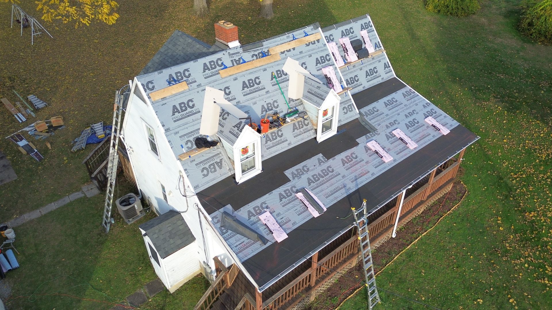 House roof under construction, covered in gray underlayment and black shingles. Ladders and tools are present.