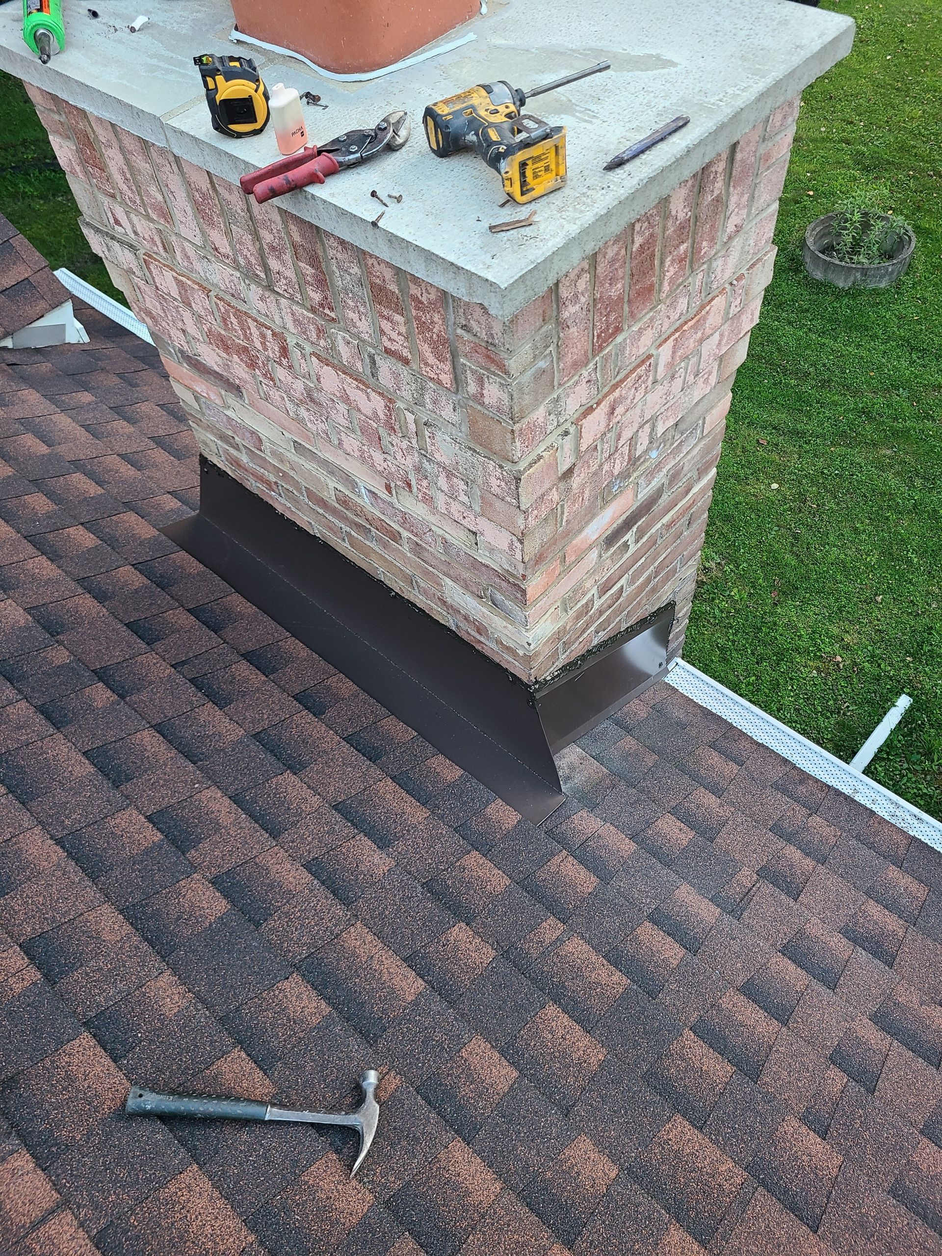 Chimney on a brown shingle roof, with tools on top. Chimney has dark flashing.