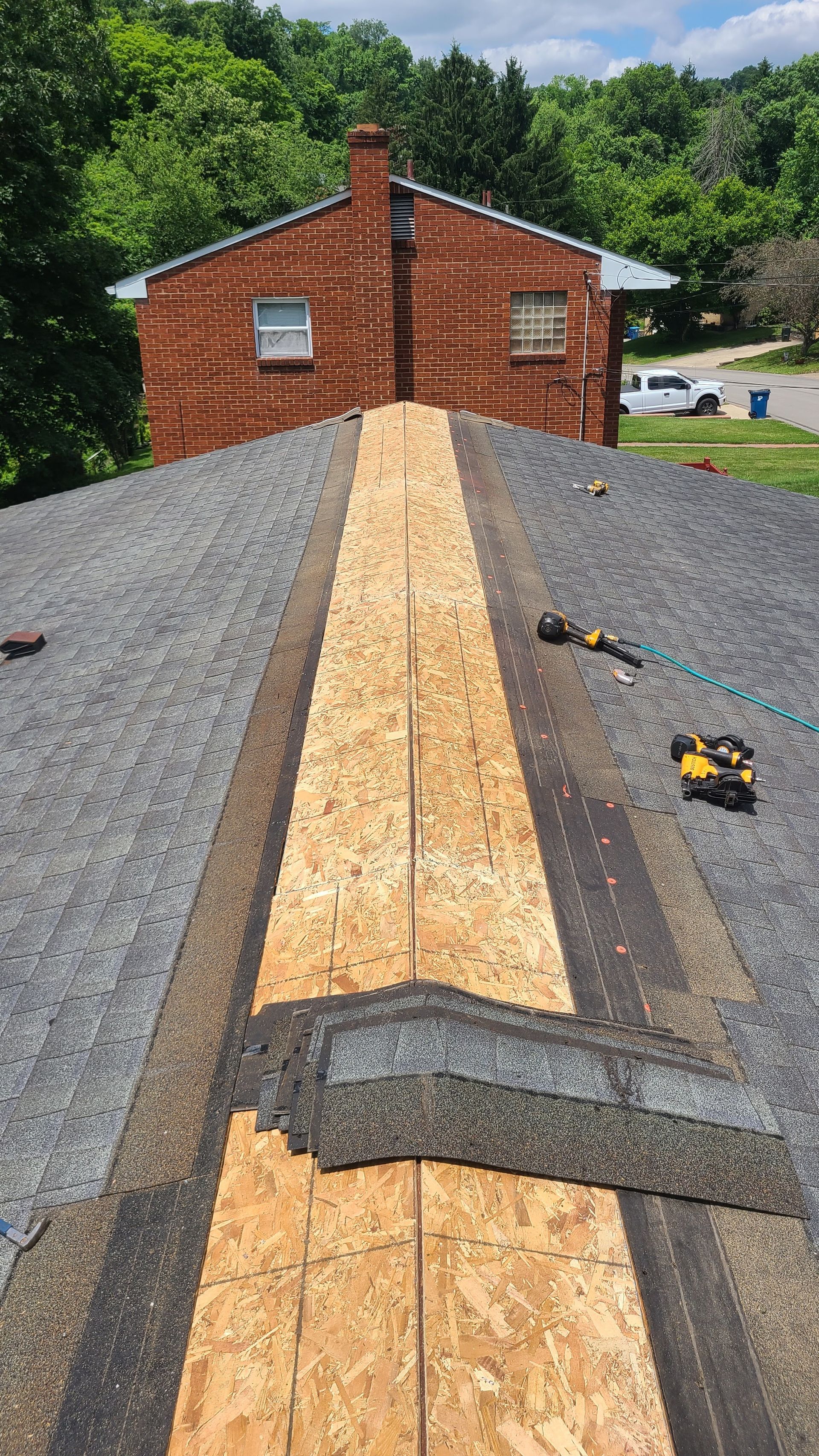 Roof with missing shingles revealing wood; brick house and green trees in the background.
