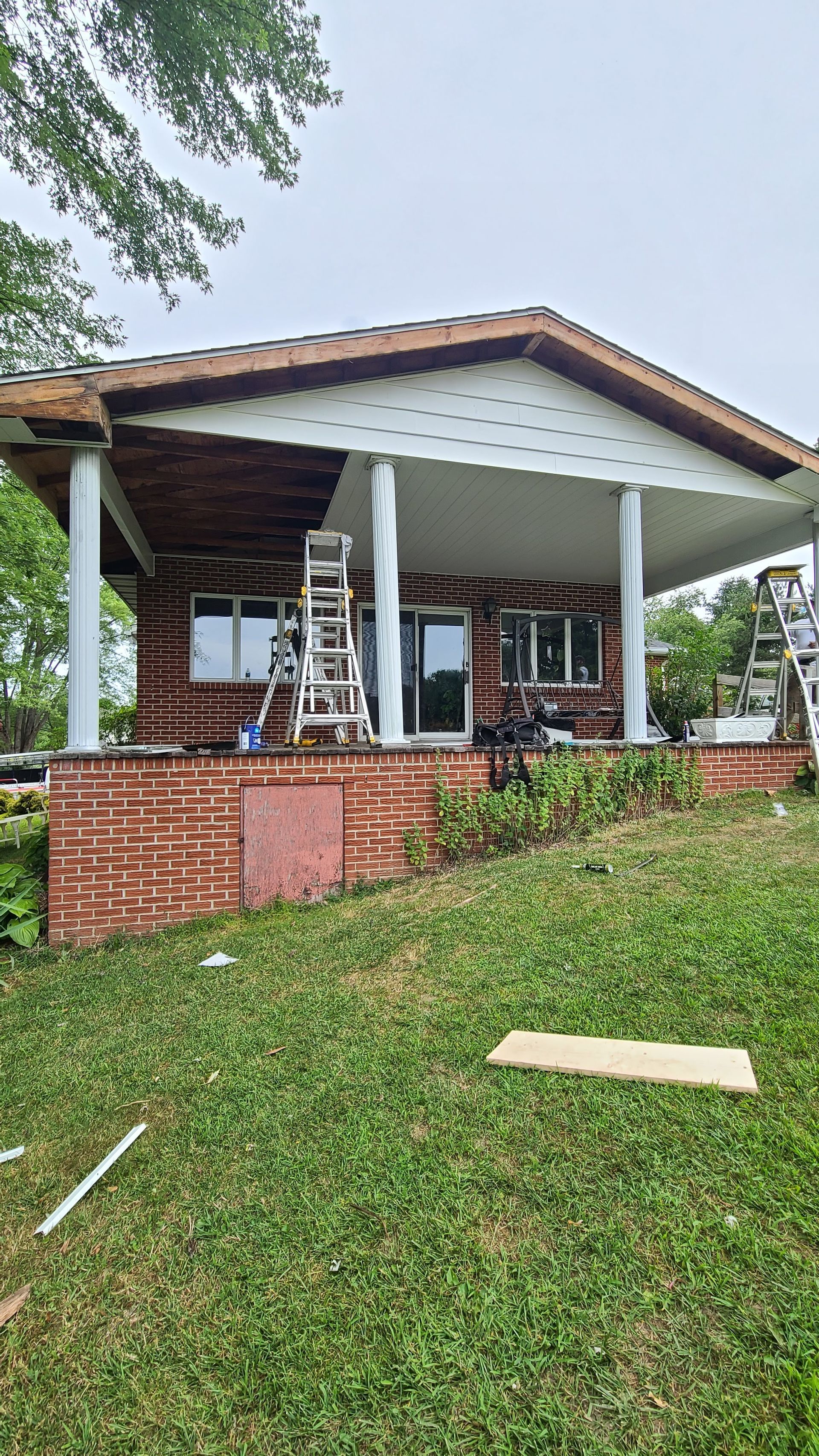 Brick house under construction; white columns, ladders, green grass, and overcast sky.