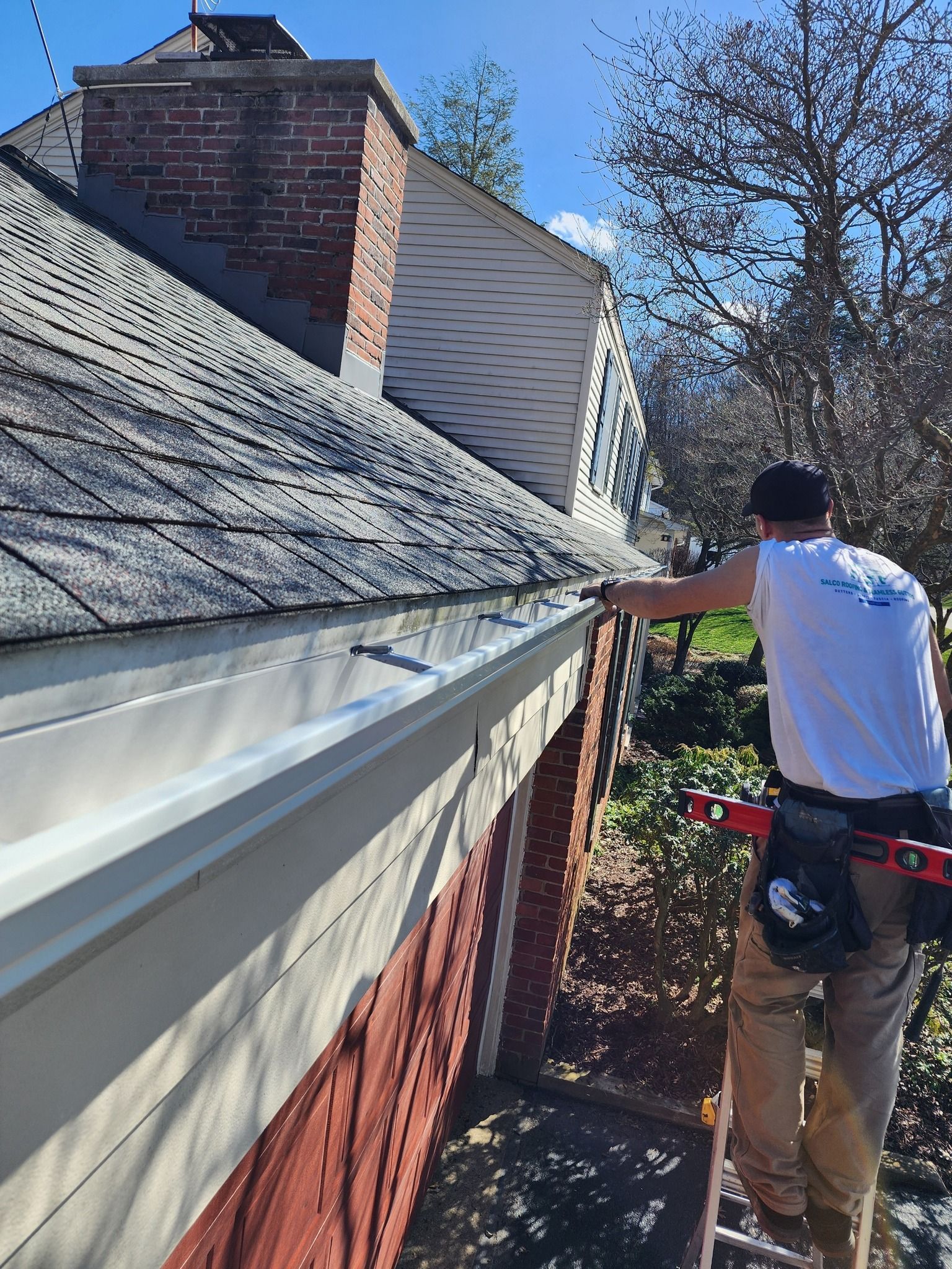 A person on a ladder cleans a roof gutter. He wears a white shirt, beige pants, and a hat. The setting is sunny.