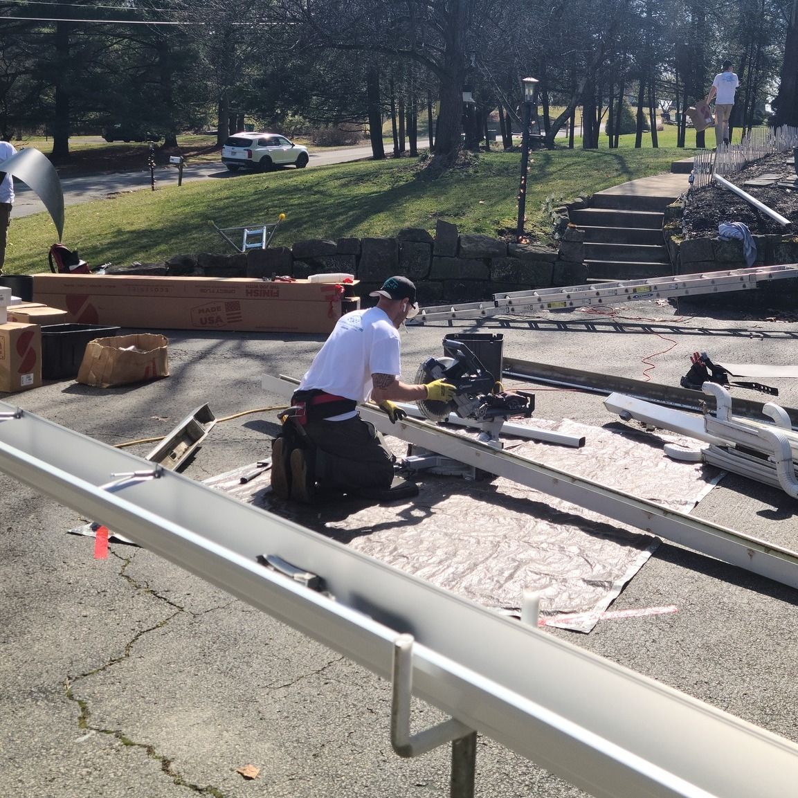 Man installing a gutter, using a saw. Outdoors with other workers and a house in the background.