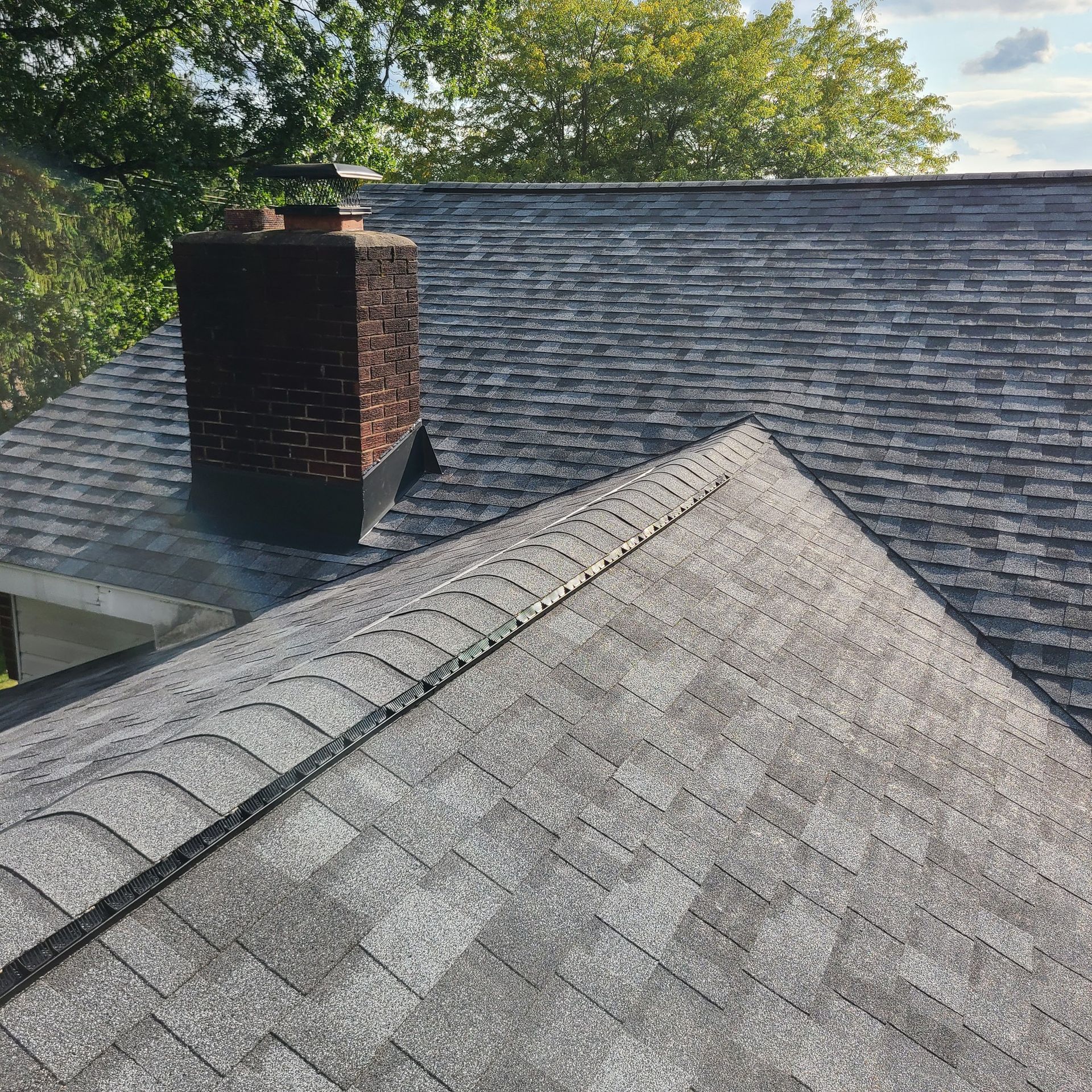Asphalt shingle roof with a brick chimney and green trees in the background.