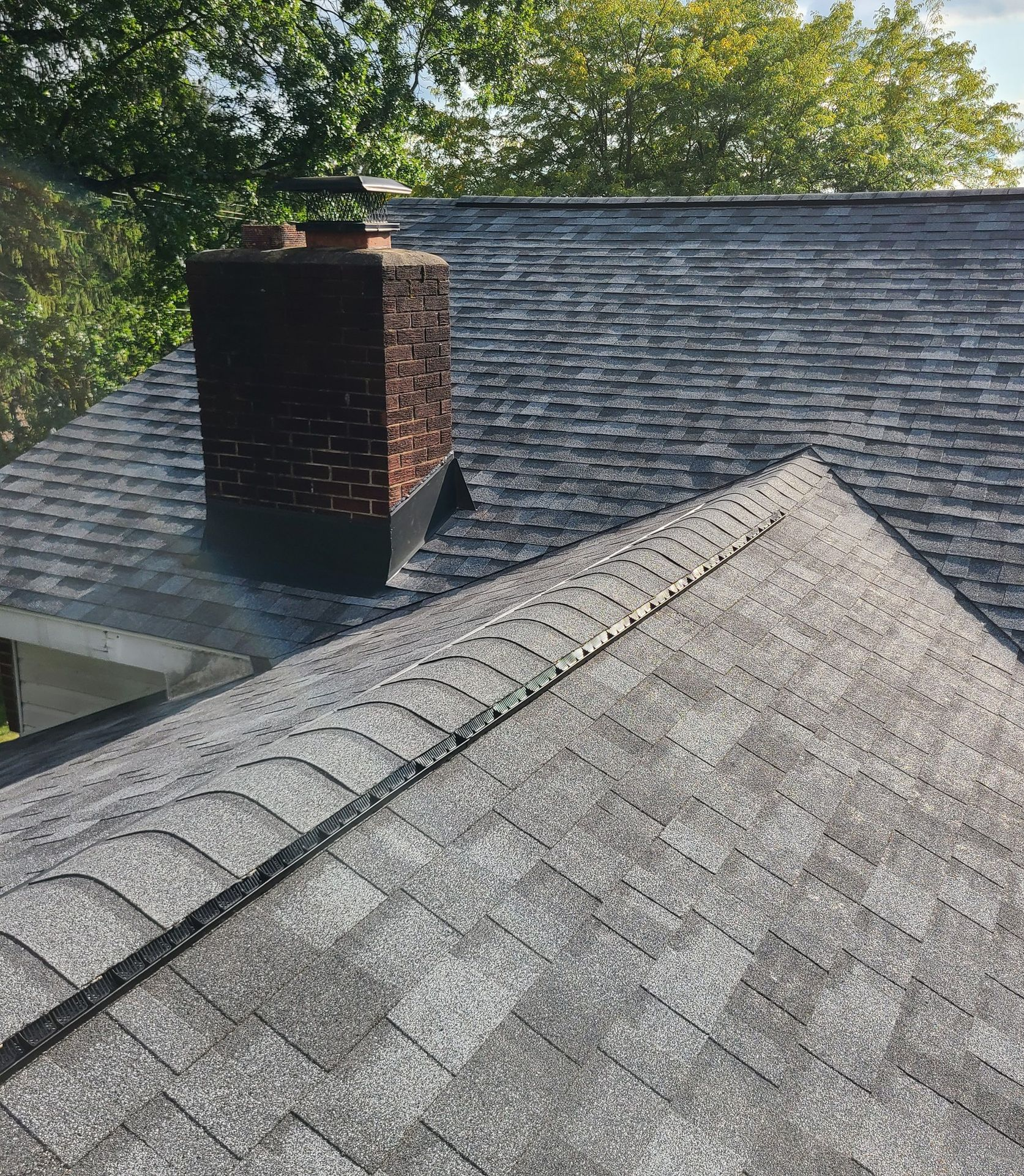 Gray asphalt shingle roof with a brick chimney and trees in the background.