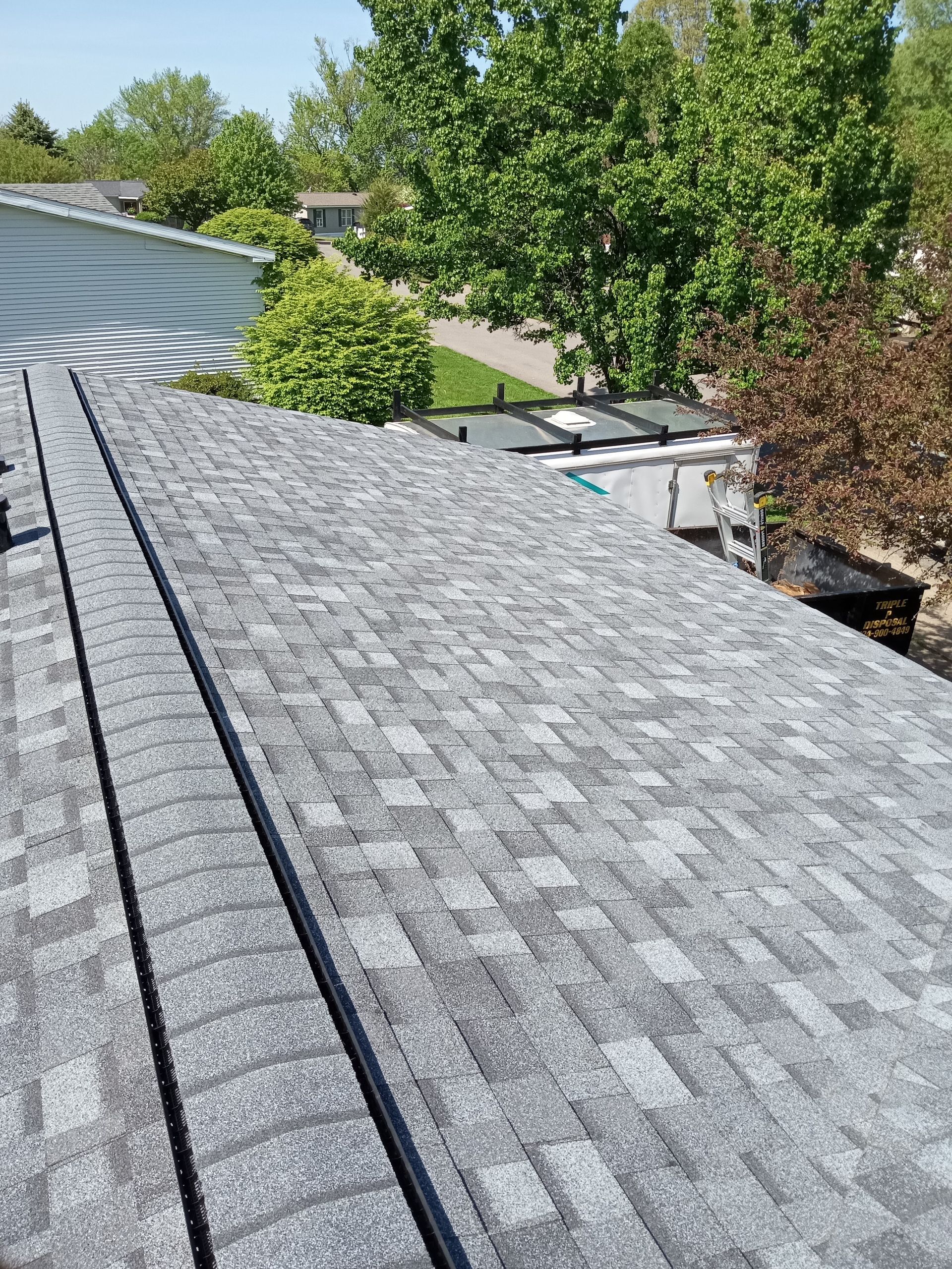 Gray asphalt shingle roof, angled view. Houses and trees in background under bright sky.