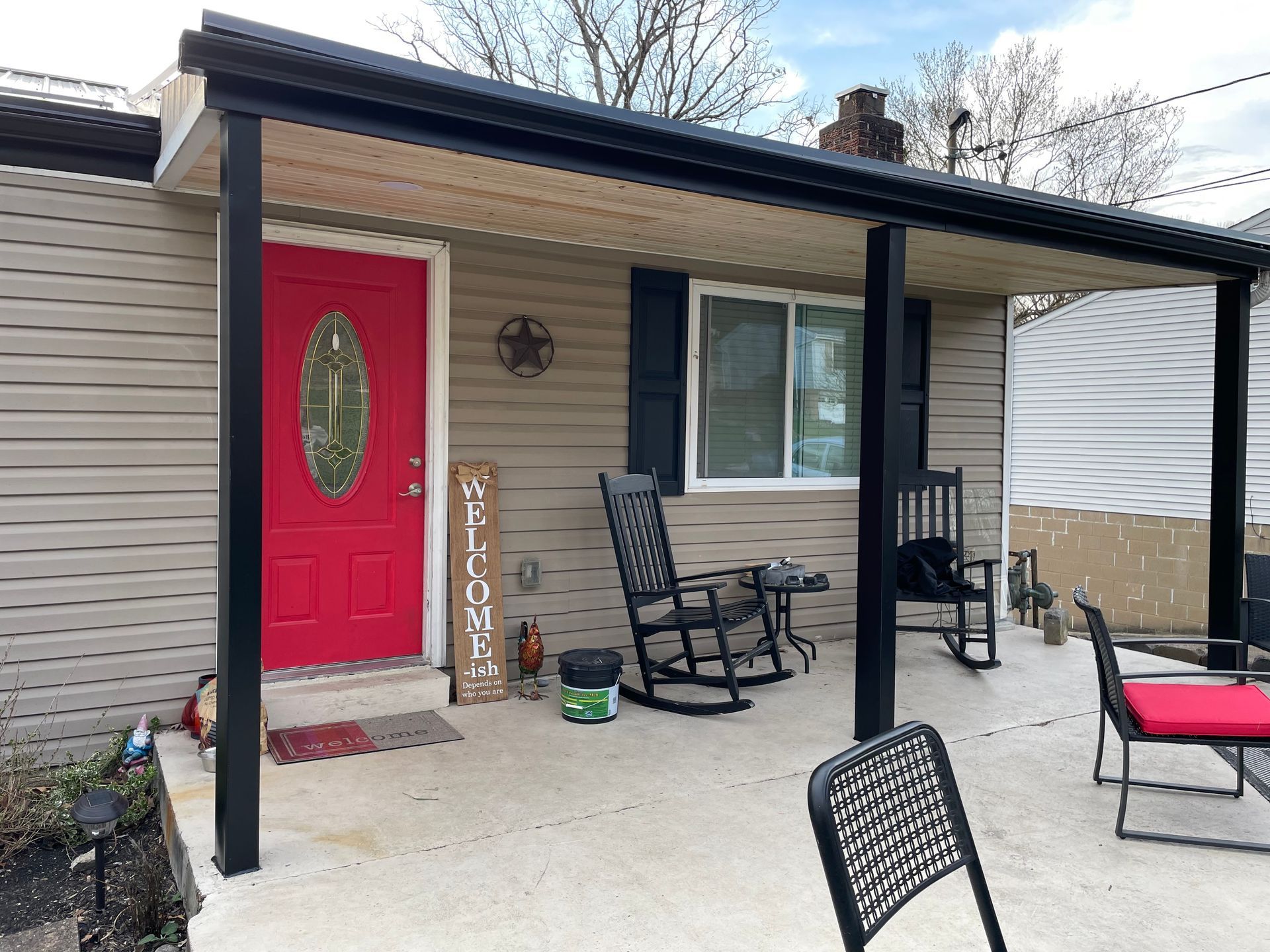 A small house with a red door and porch, with black framing. Two rocking chairs and outdoor seating.