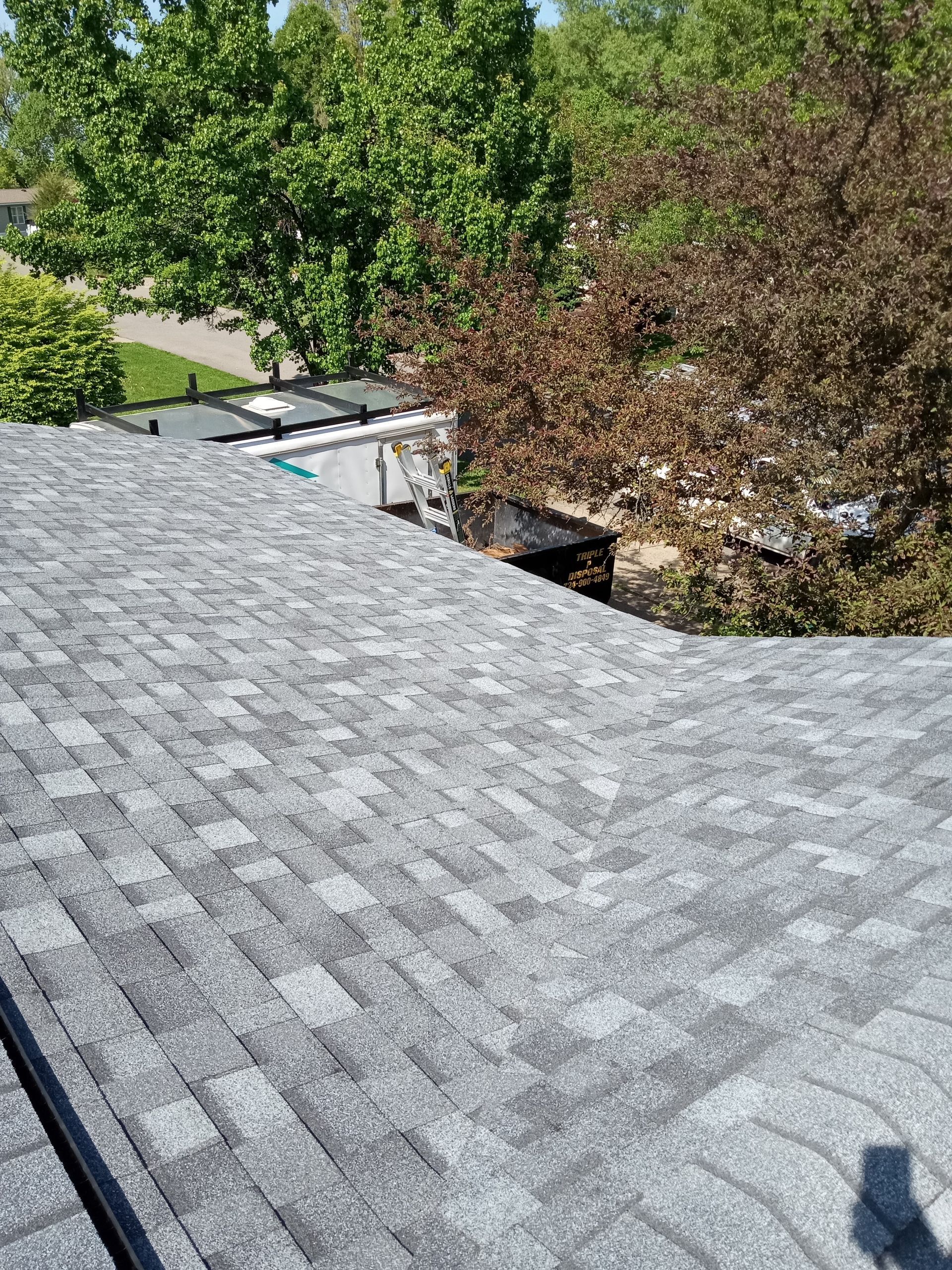 Gray asphalt shingle roof with a damaged section, surrounded by trees and a sunny sky.