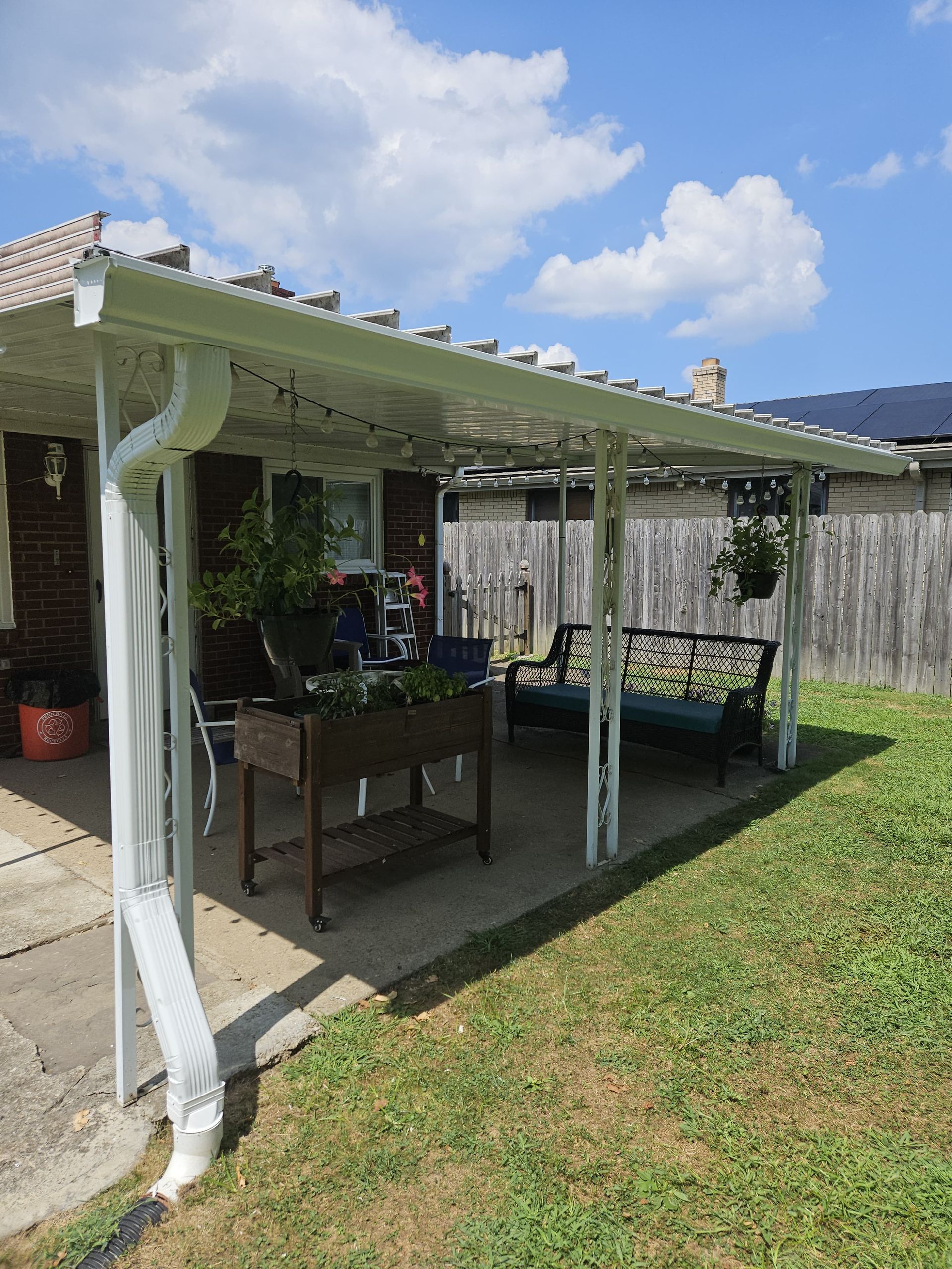 White-framed patio cover over concrete with a bench, table with plants, and a grassy yard.