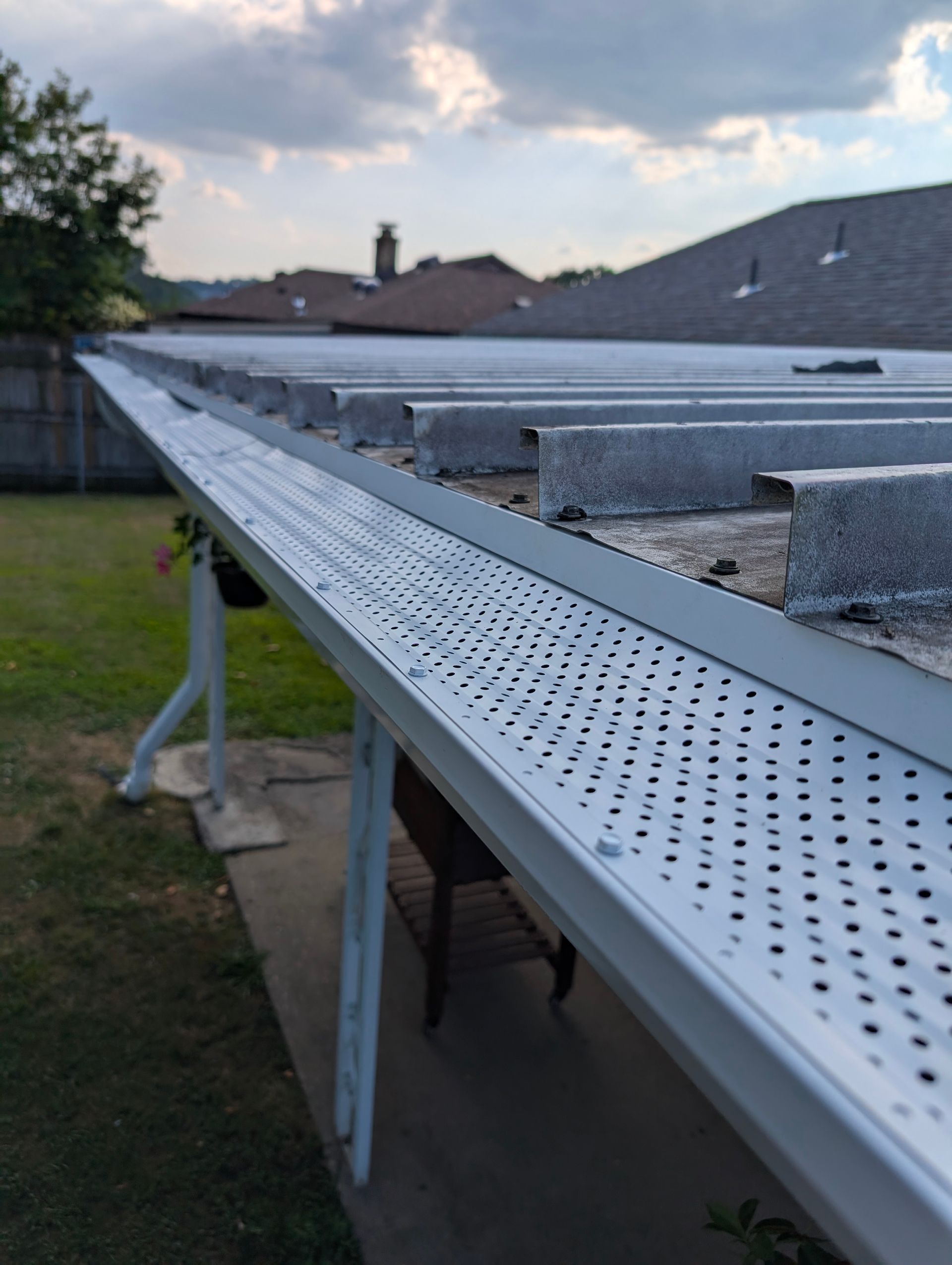 Silver gutter guard on a roof with visible perforations. Cloudy sky in the background, green grass and a house.