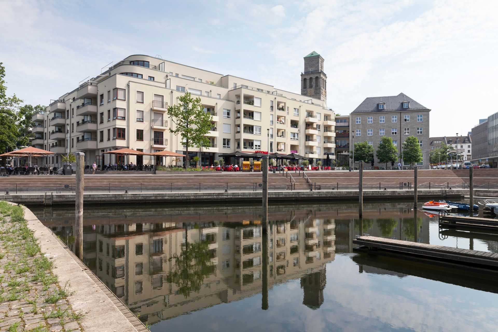 Moderne Apartments spiegeln sich im ruhigen Wasser eines Hafens, im Hintergrund ist ein Kirchturm zu sehen.