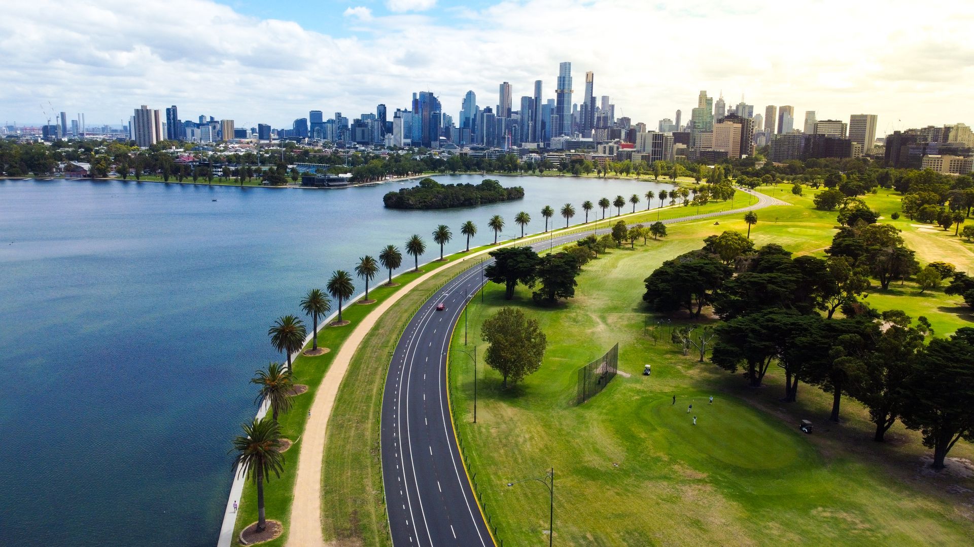Albert Park Lake, Melbourne City, Victoria, Australia. Aerial drone view of the lake with a row of palm trees, the road, the open-wheel single-seater racing car grand prix racing track, walking track and bike riders. Sky-rise buildings and the Melbourne skyline in the background.