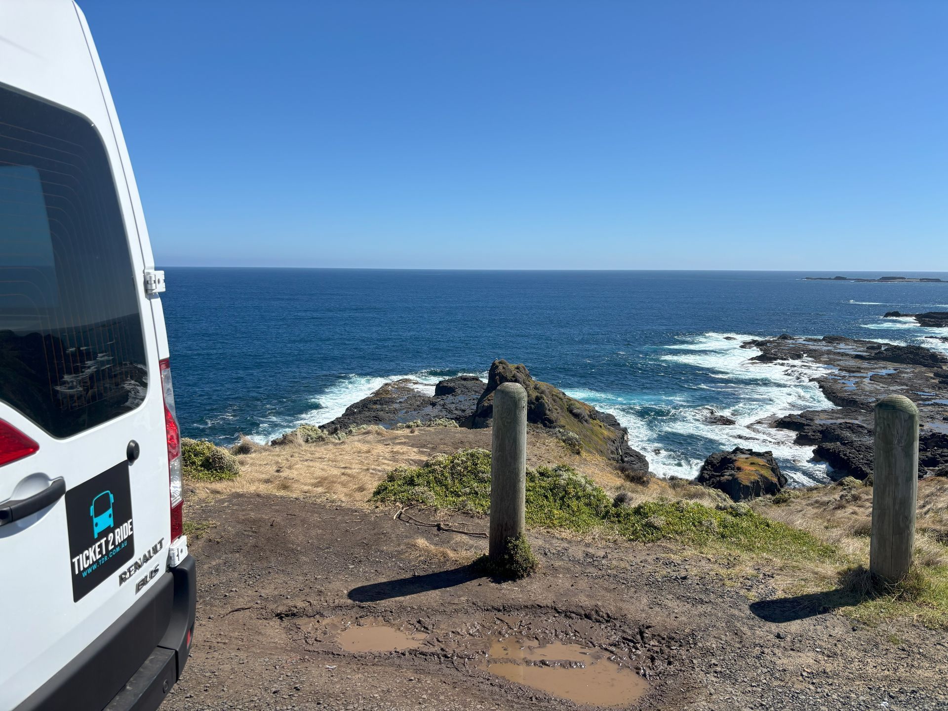 Ticket 2 Ride minibus parked overlooking ocean at Phillip Island. Rocky coast, blue water, bright sunny day.