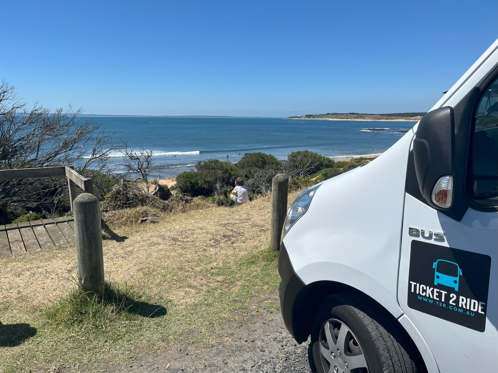 White minibus parked by a beach, Ticket 2 Ride logo, person near water, blue sky.