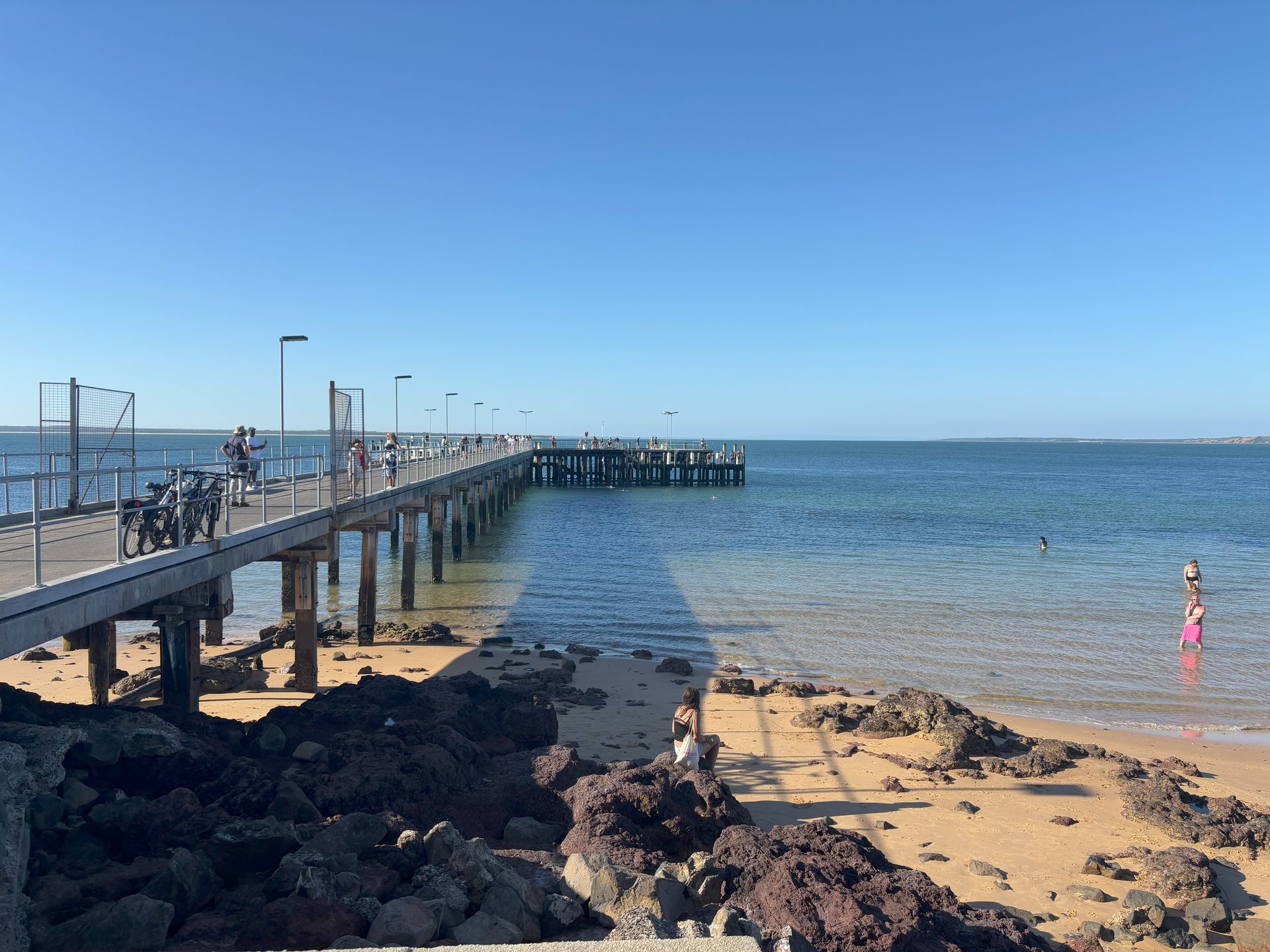 Pier extending over clear water, with people and bikes. Sunny day at the beach.