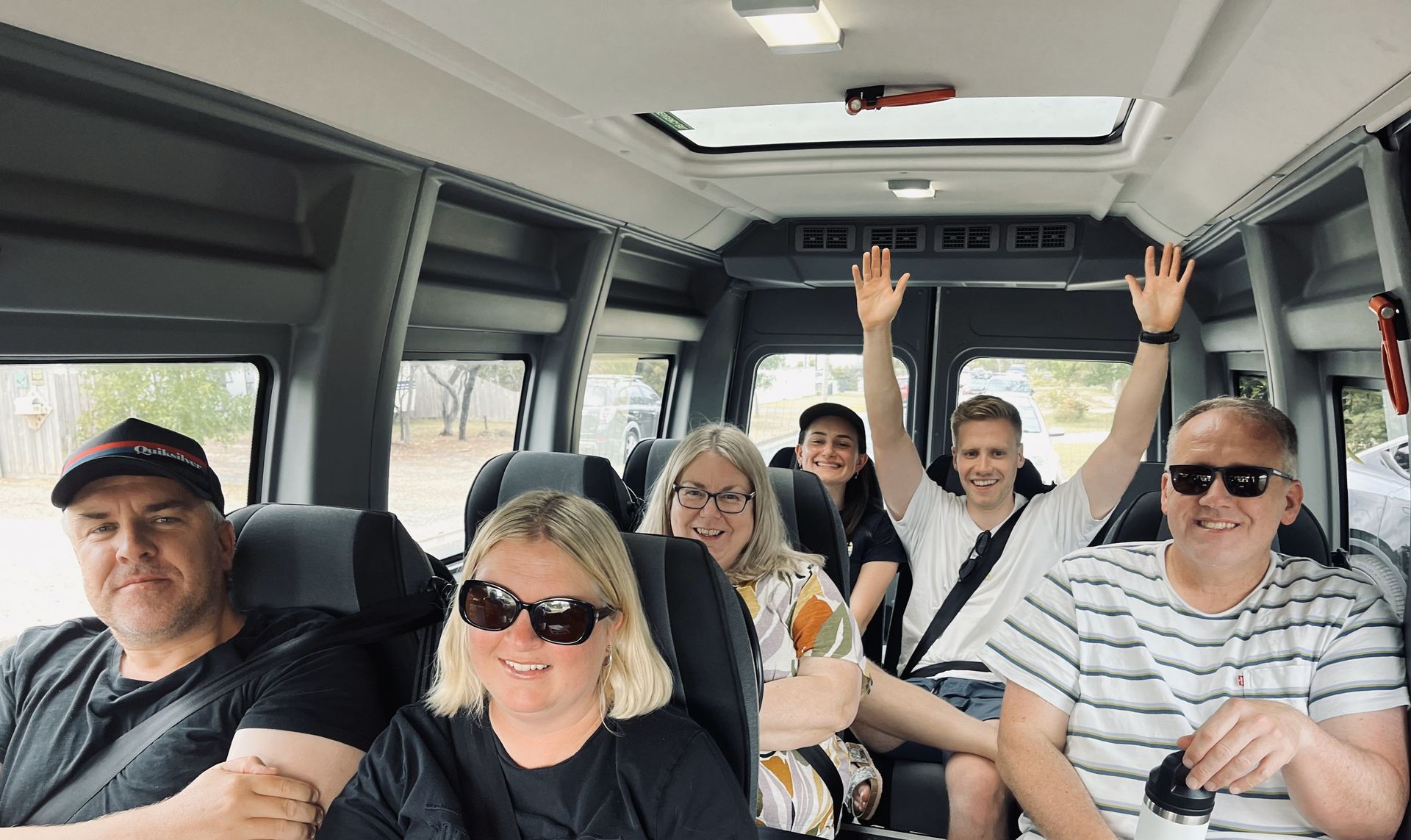 People in a Ticket 2 Ride minibus, some smiling, waving, and wearing sunglasses, enjoying a trip together.