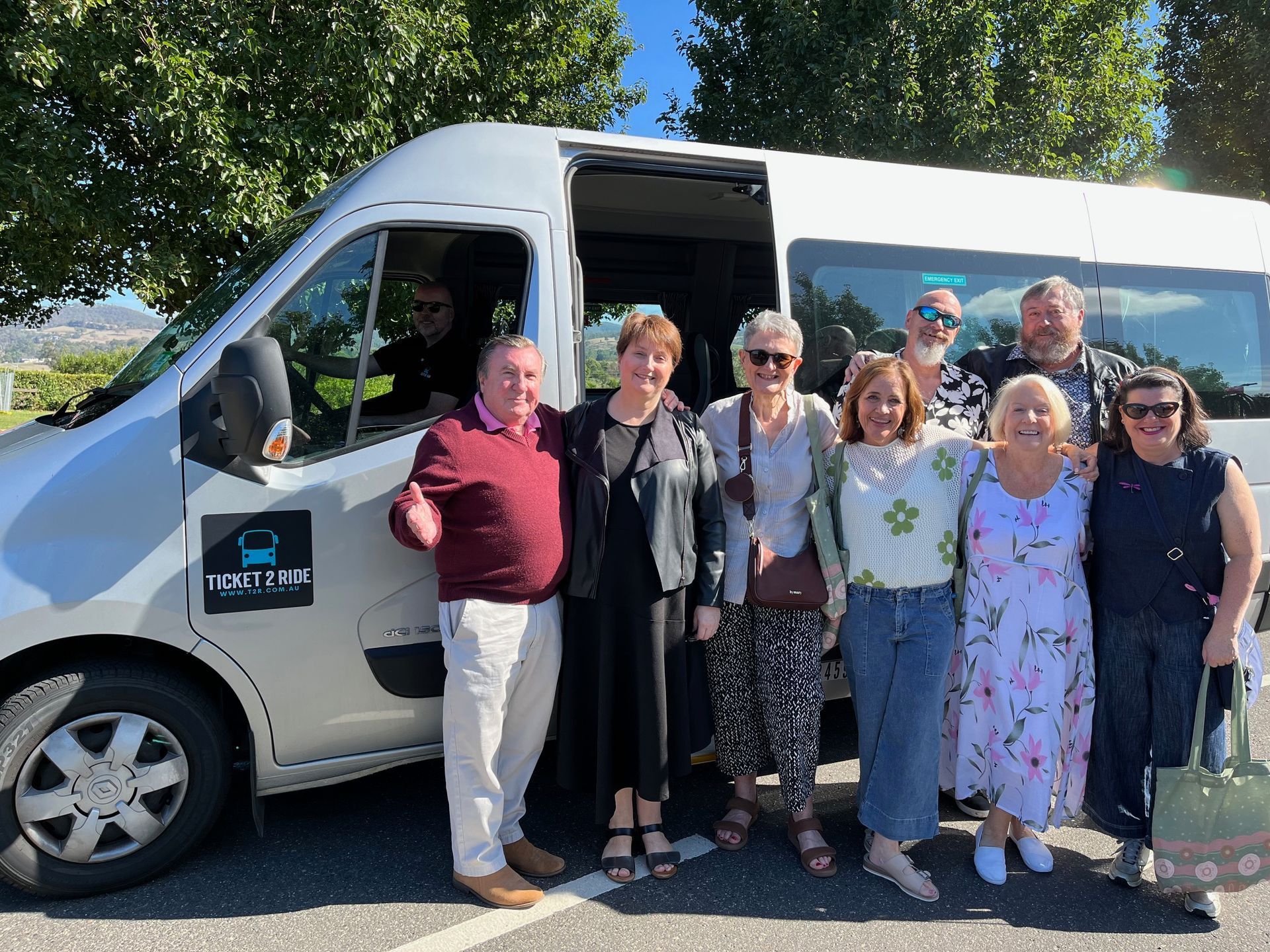 Group of people posing with a silver Ticket 2 Ride minibus. Man gives thumbs up. Sunny outdoors.