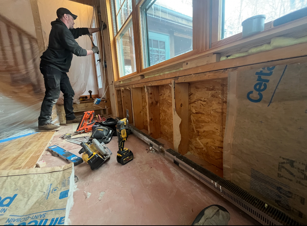 Construction worker installing wiring near a window. Interior of a building with exposed framing and tools.