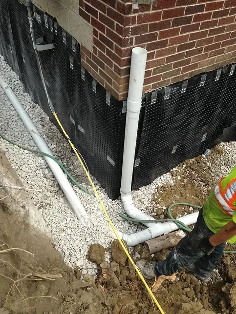 A man is measuring a pipe in the dirt in front of a brick building.