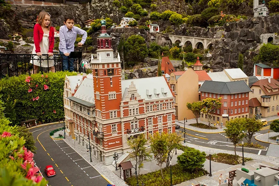 Uma criança e um adulto observam uma maquete em miniatura de uma cidade europeia de tijolos vermelhos, com ruas e áreas verdes.