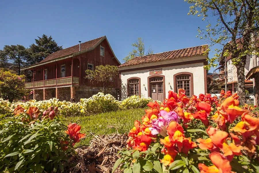 Um jardim com flores laranja brilhantes em primeiro plano, de frente para dois edifícios rústicos e antiquados sob um céu azul claro.