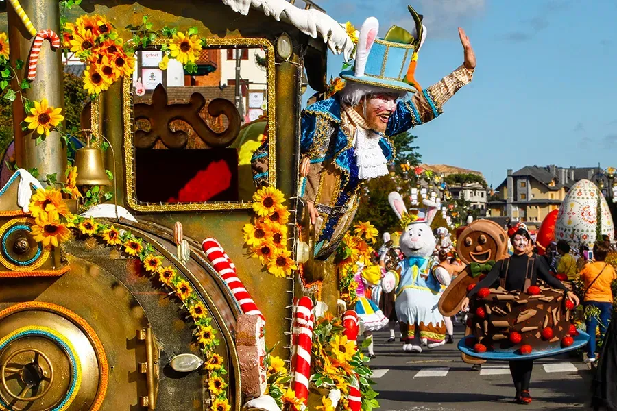 Um carro alegórico decorado com girassóis apresenta uma pessoa fantasiada de Chapeleiro Maluco acenando perto de artistas de rua.