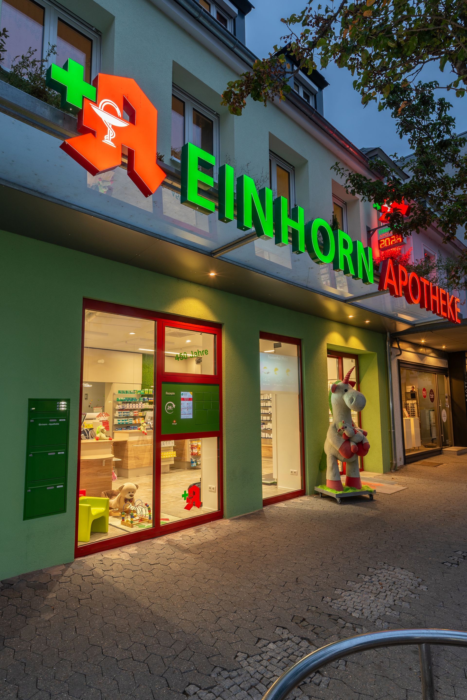 Grüne Fassade der Einhorn Apotheke mit rot-grünem Schild, Glastüren und einer kleinen Hundestatue.