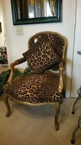 Leopard print upholstered chair with gold trim and a matching pillow, against a white wall.