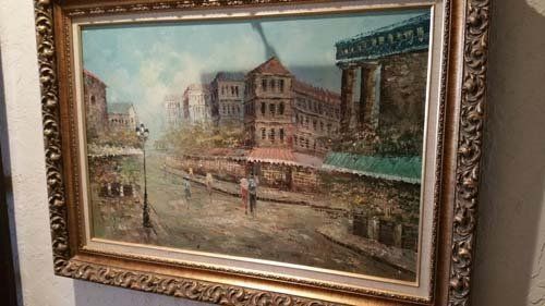 Oil painting of a European street scene with buildings and figures under a cloudy sky, in a gold ornate frame.