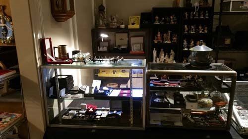 Display cases in a shop filled with various items like watches, clocks, and figurines. Dark interior, lit displays.