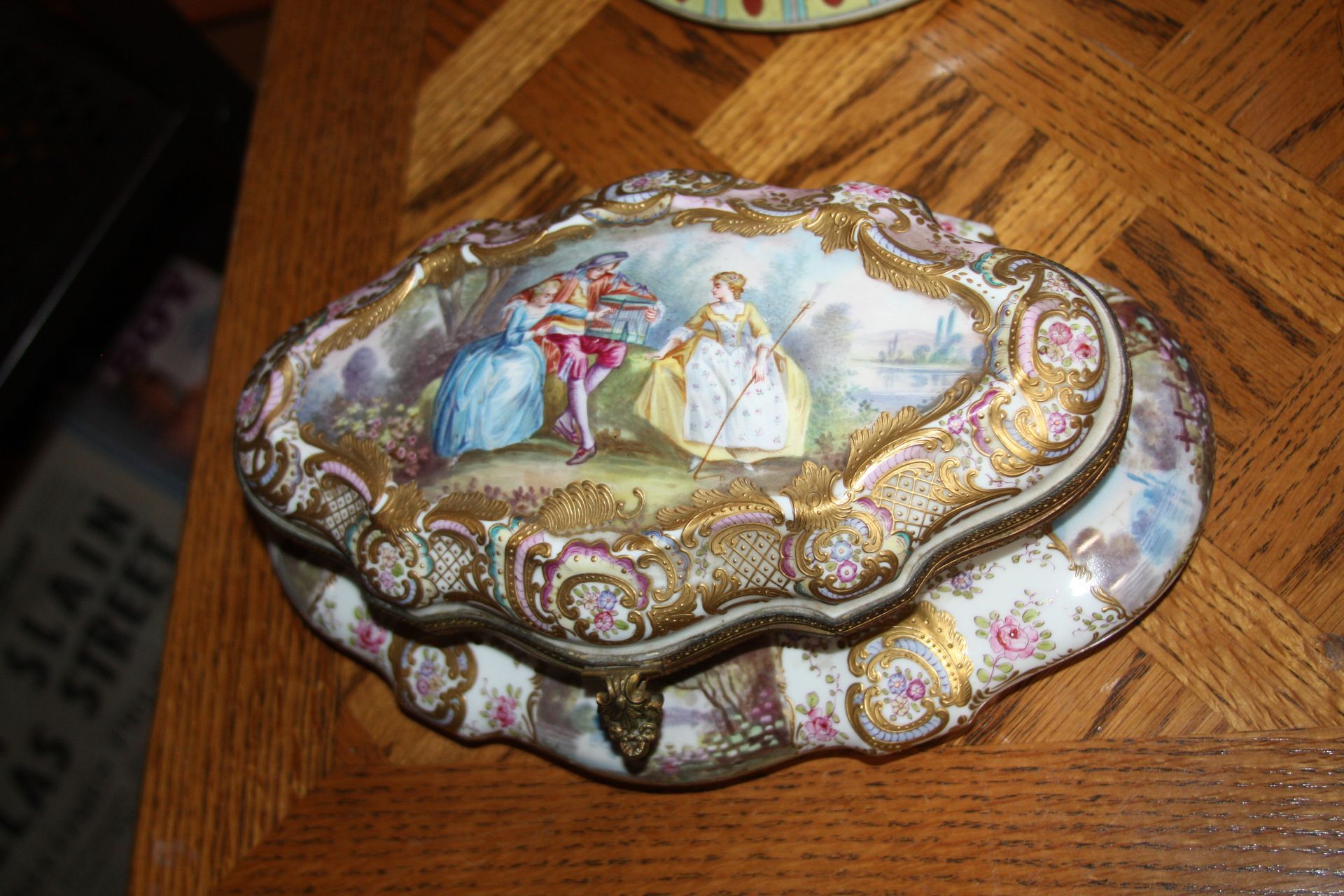 Antique porcelain box with hand-painted scene of figures and floral details, gold trim, on wooden surface.