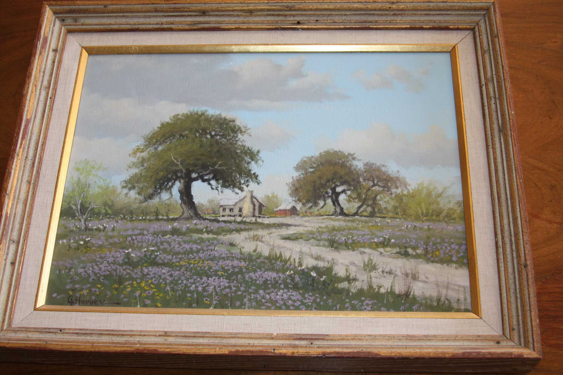 Landscape painting of bluebonnets, trees, dirt road, and small buildings under a cloudy sky.