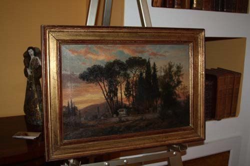 Oil painting of a landscape with trees, framed in gold, displayed on an easel. Sunset hues.