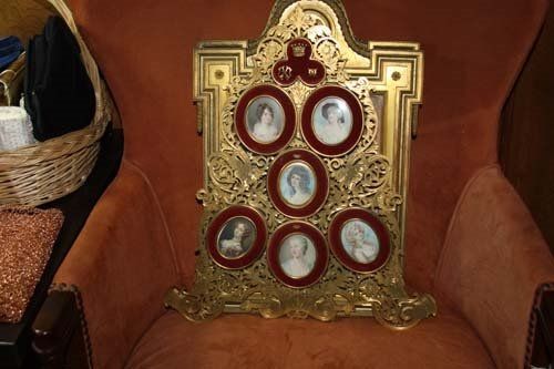 Ornate golden framed display with seven portrait miniatures, on an orange armchair.