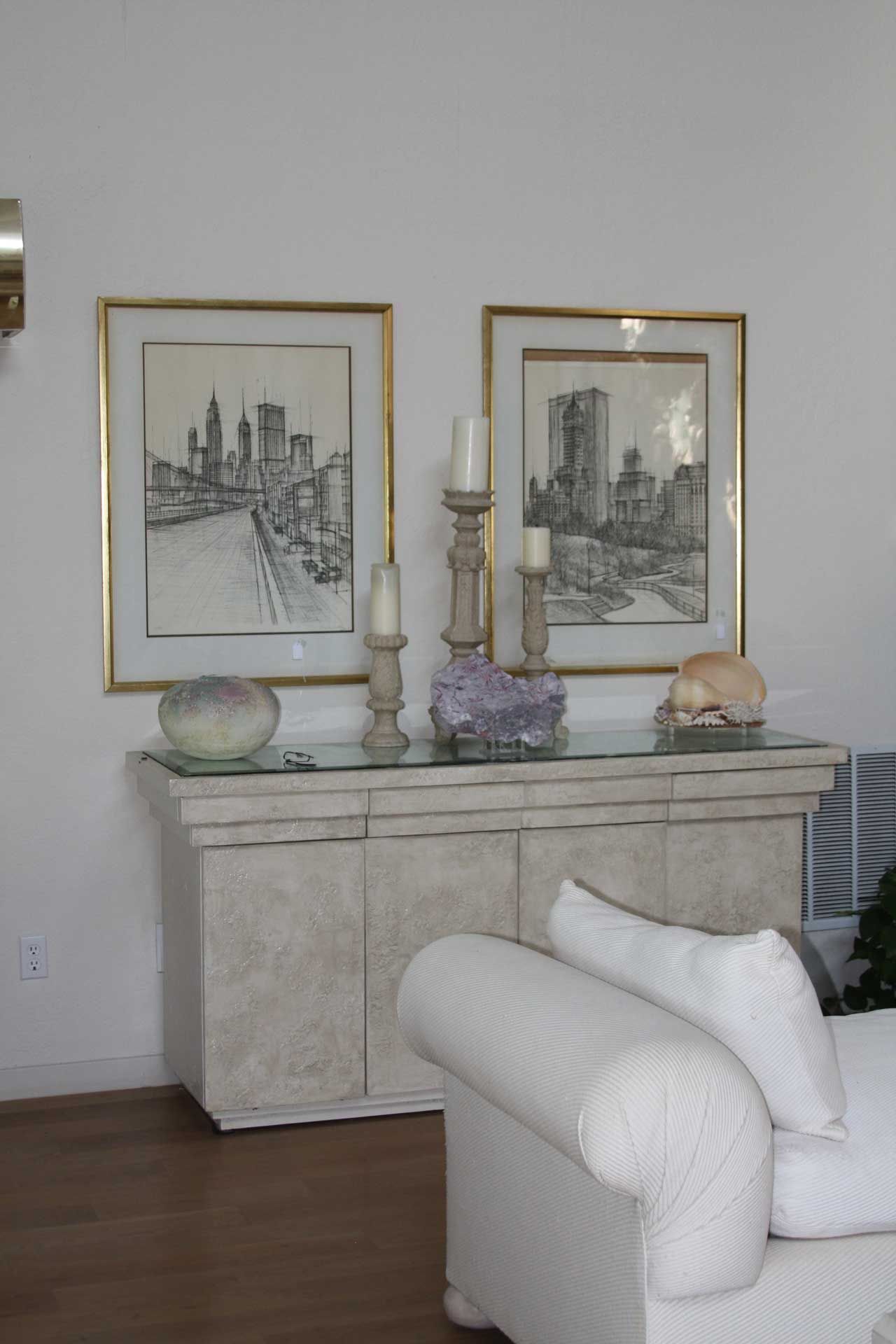 A white credenza with two framed city skyline drawings and candles, next to a white couch.