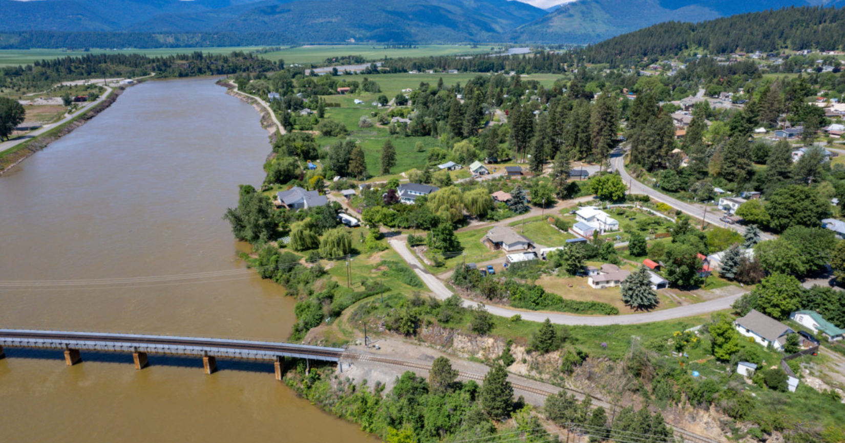 Aerial view of a town next to a muddy river, with a bridge in the foreground and mountains in the background.