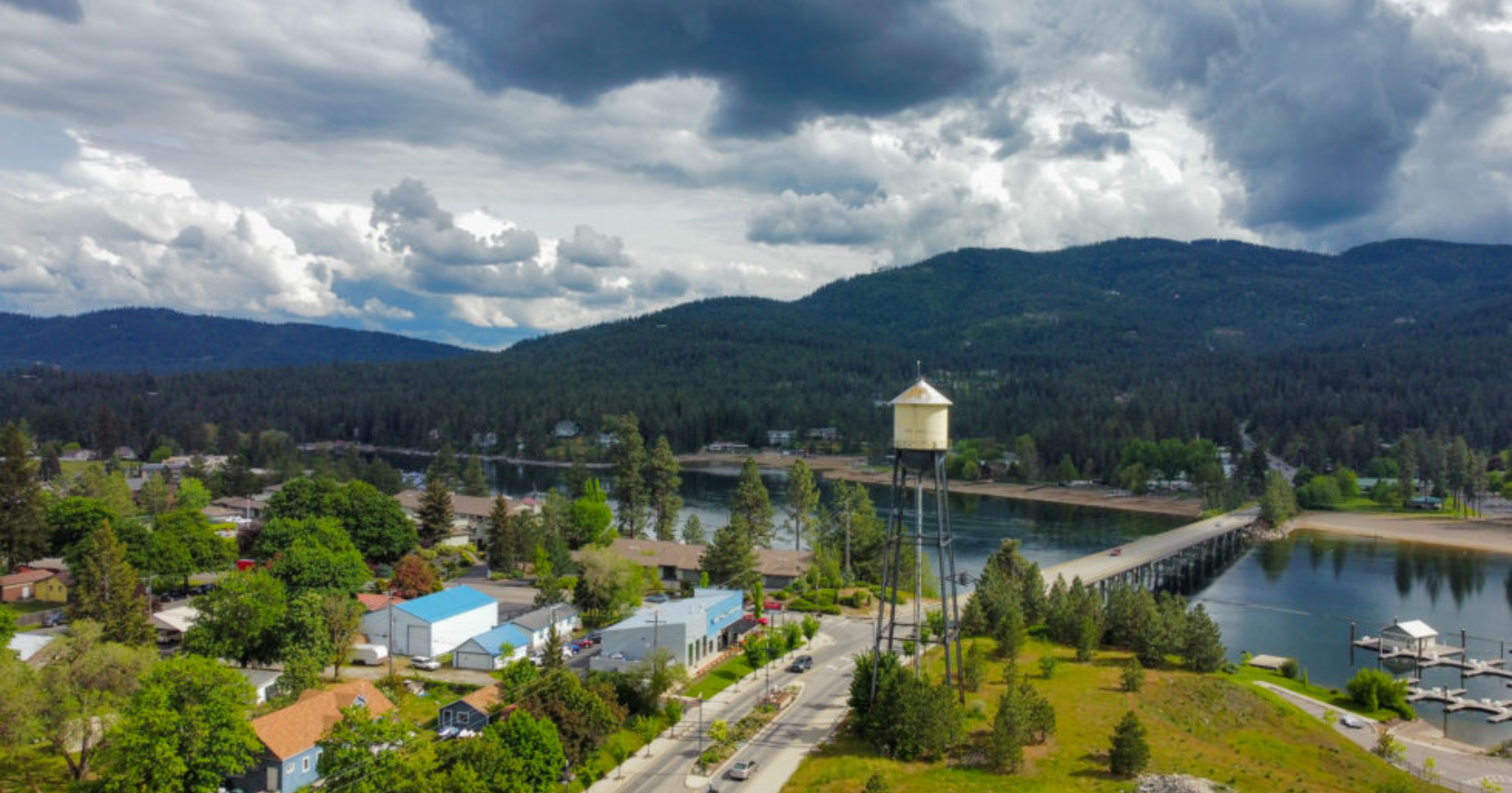 Aerial view of a town with a lake, bridge, and mountains under a cloudy sky. A water tower stands tall.
