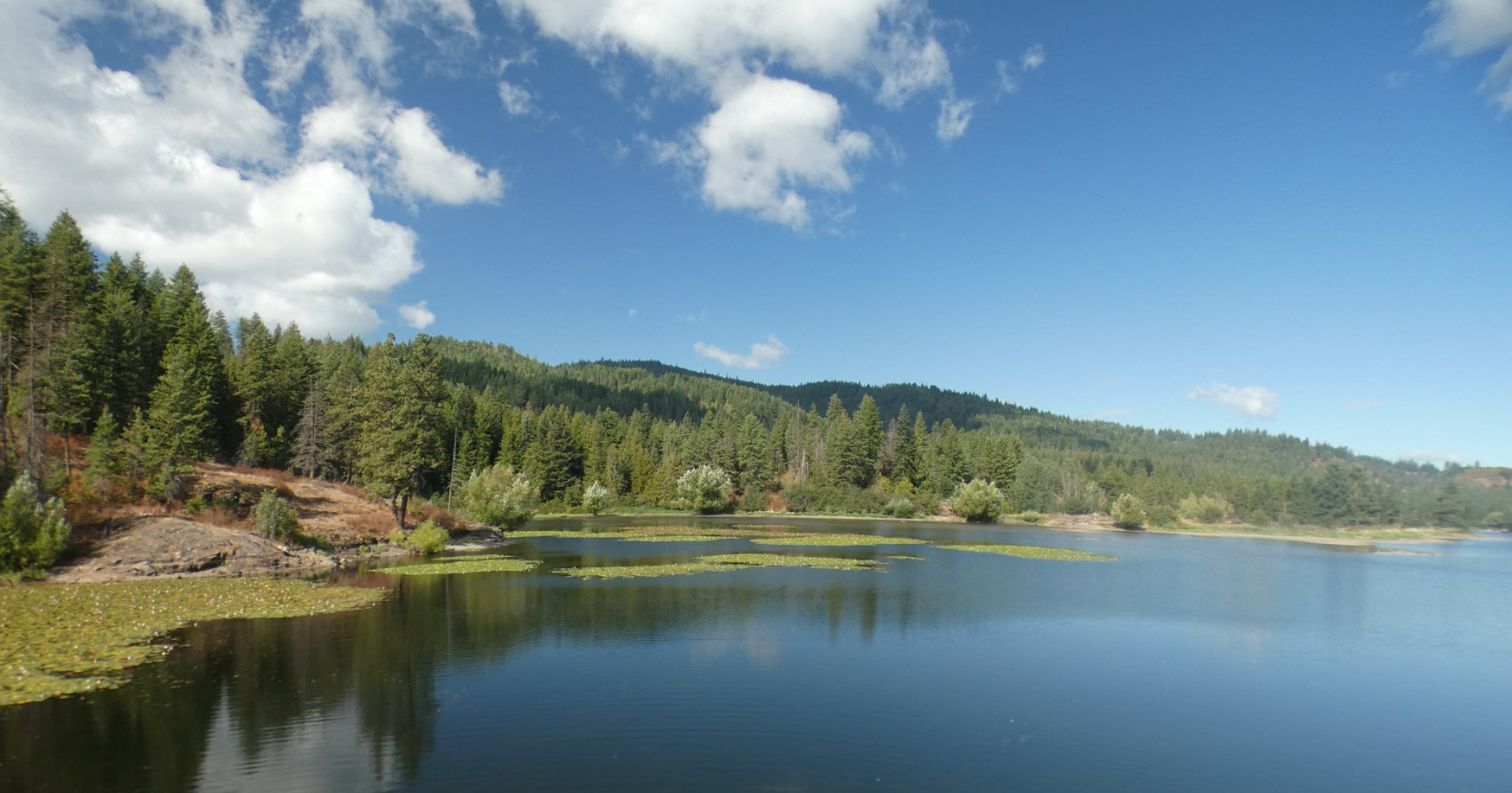 Lake with blue water reflecting the sky, surrounded by green trees and hills under a partly cloudy sky.