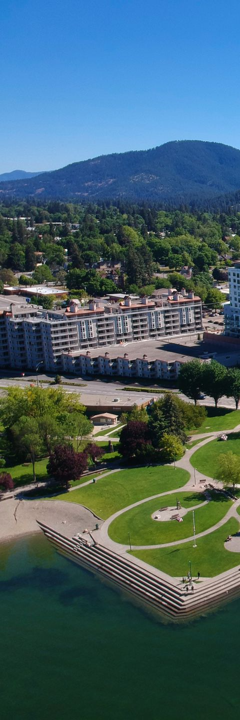 Aerial view of park and buildings along waterfront with mountains in the background under blue sky.