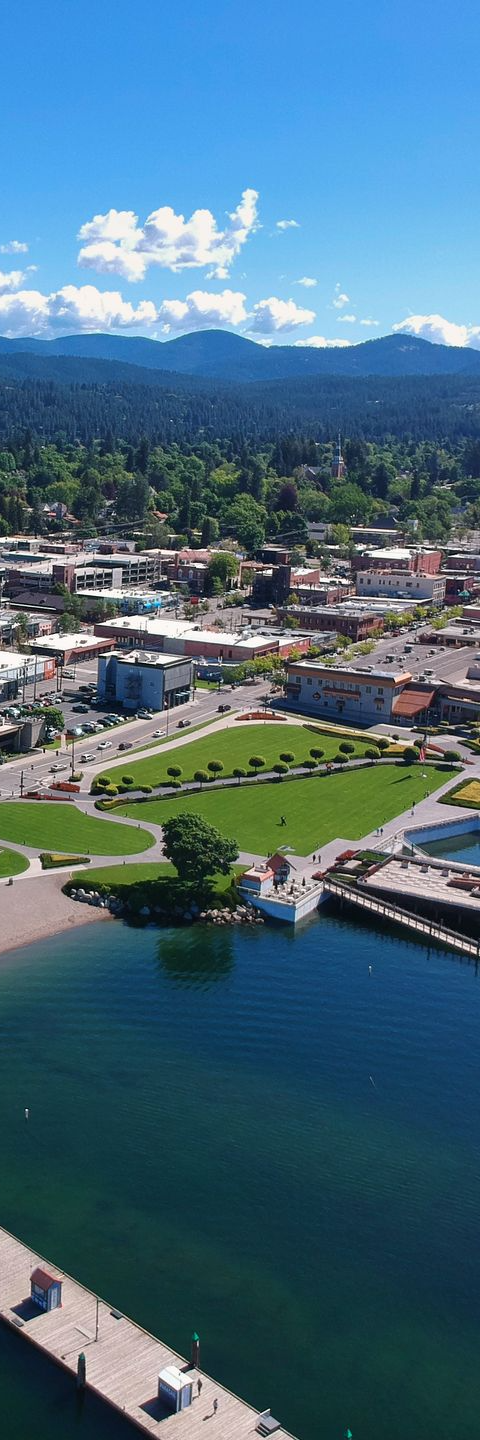 Aerial view of a city on a lake with a park, buildings, and mountains in the background under a blue sky.