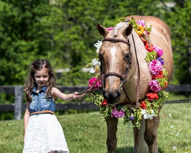 A little girl is holding the reins of a horse with flowers on its head.