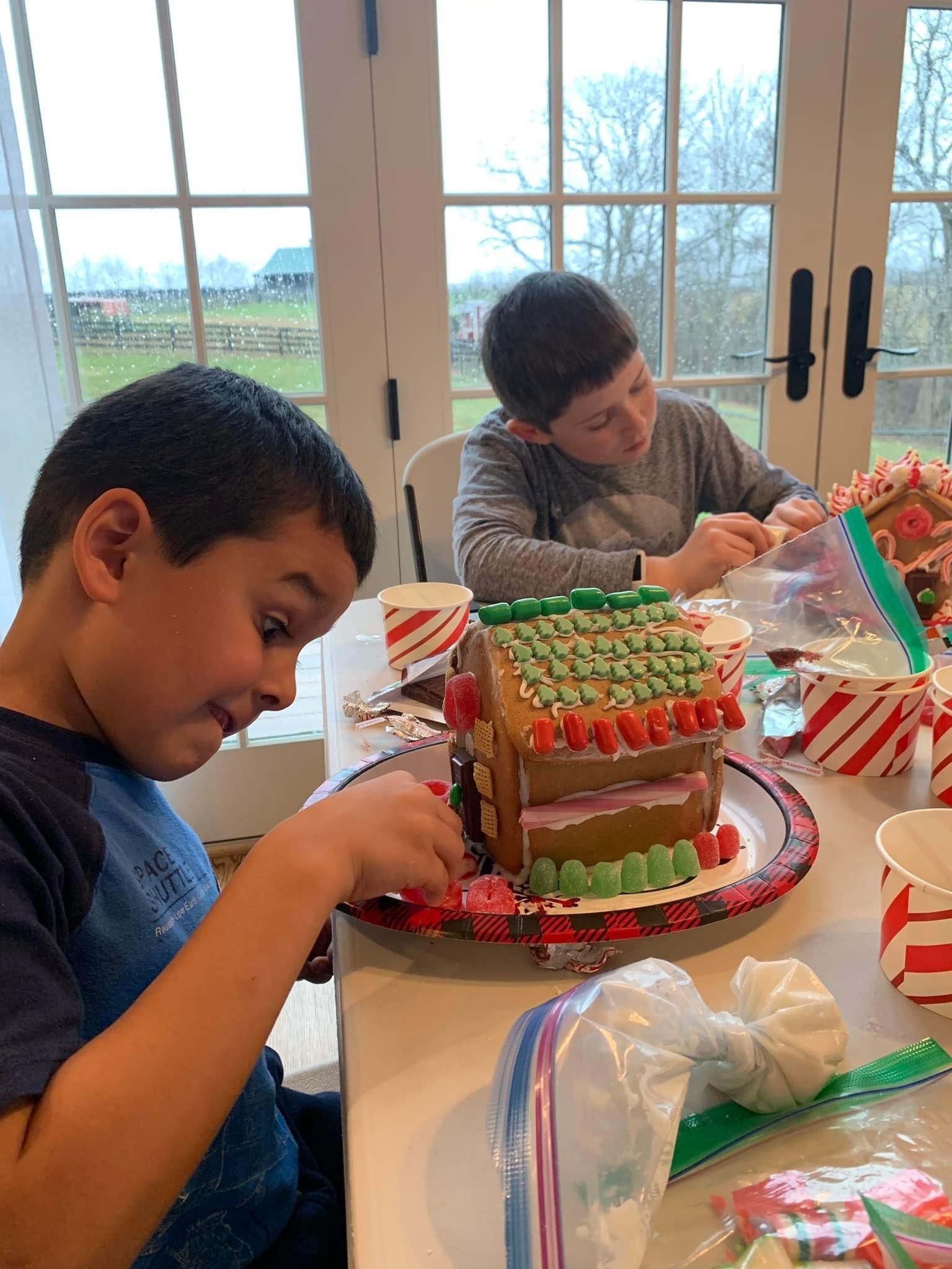 Two young boys are decorating a gingerbread house at a table.