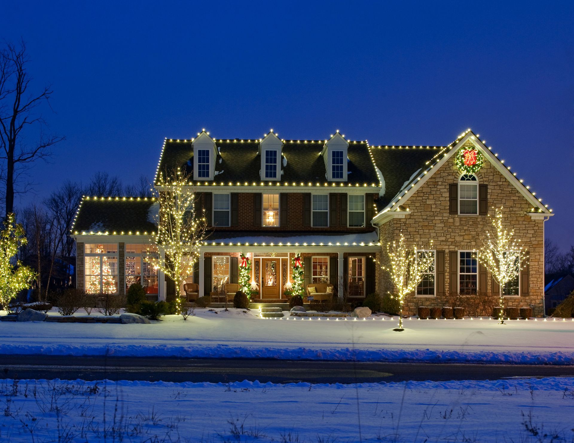 House lit with white Christmas lights and holiday decorations, snow-covered lawn.
