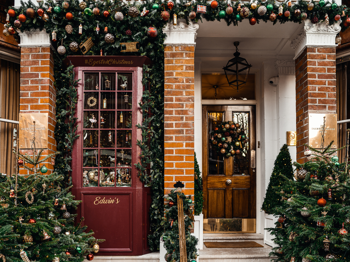 Two decorated doorways, red and wooden, adorned with Christmas garlands and trees, brick facade.