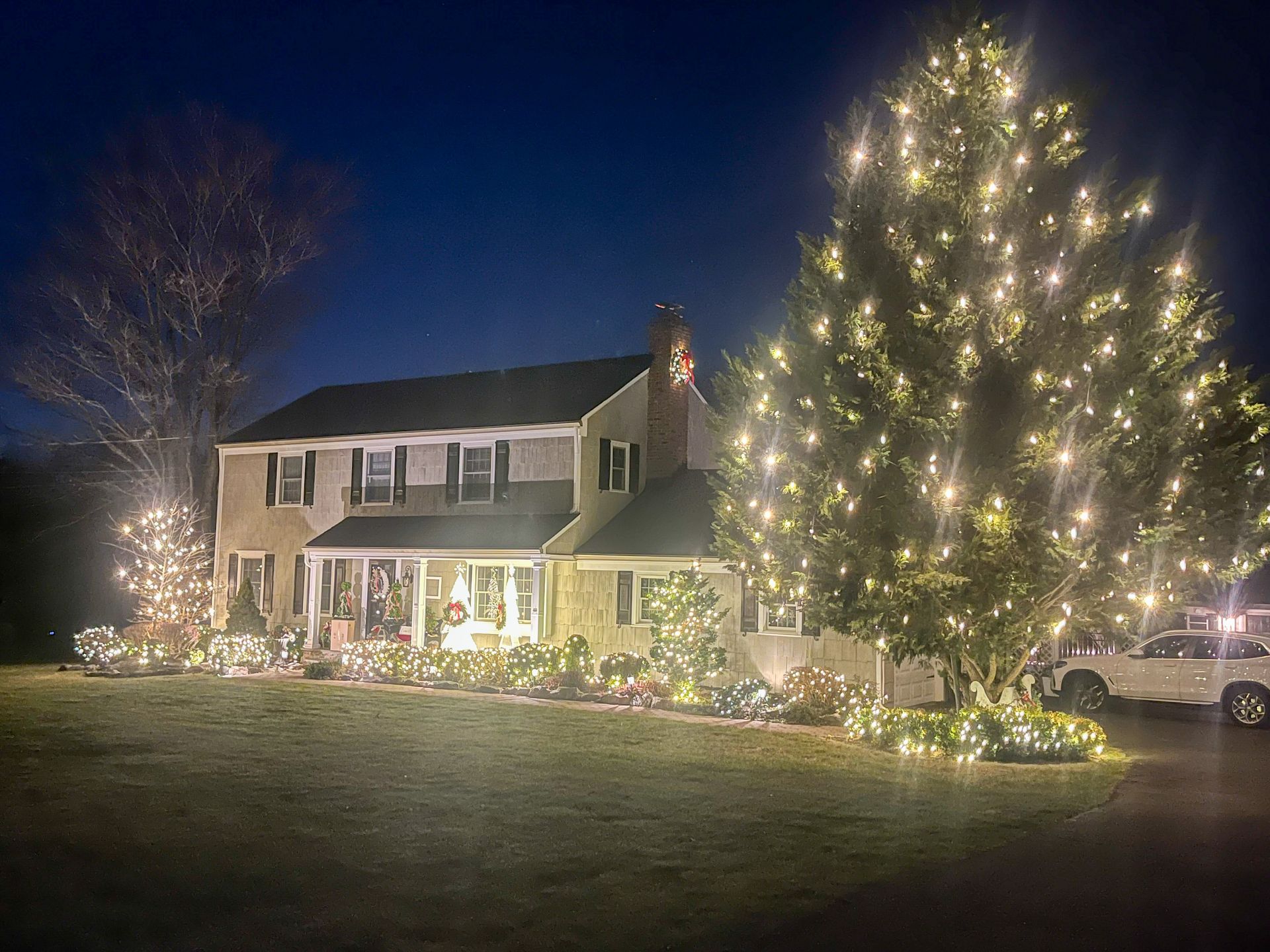 House decorated with Christmas lights at night; trees and bushes also lit up.