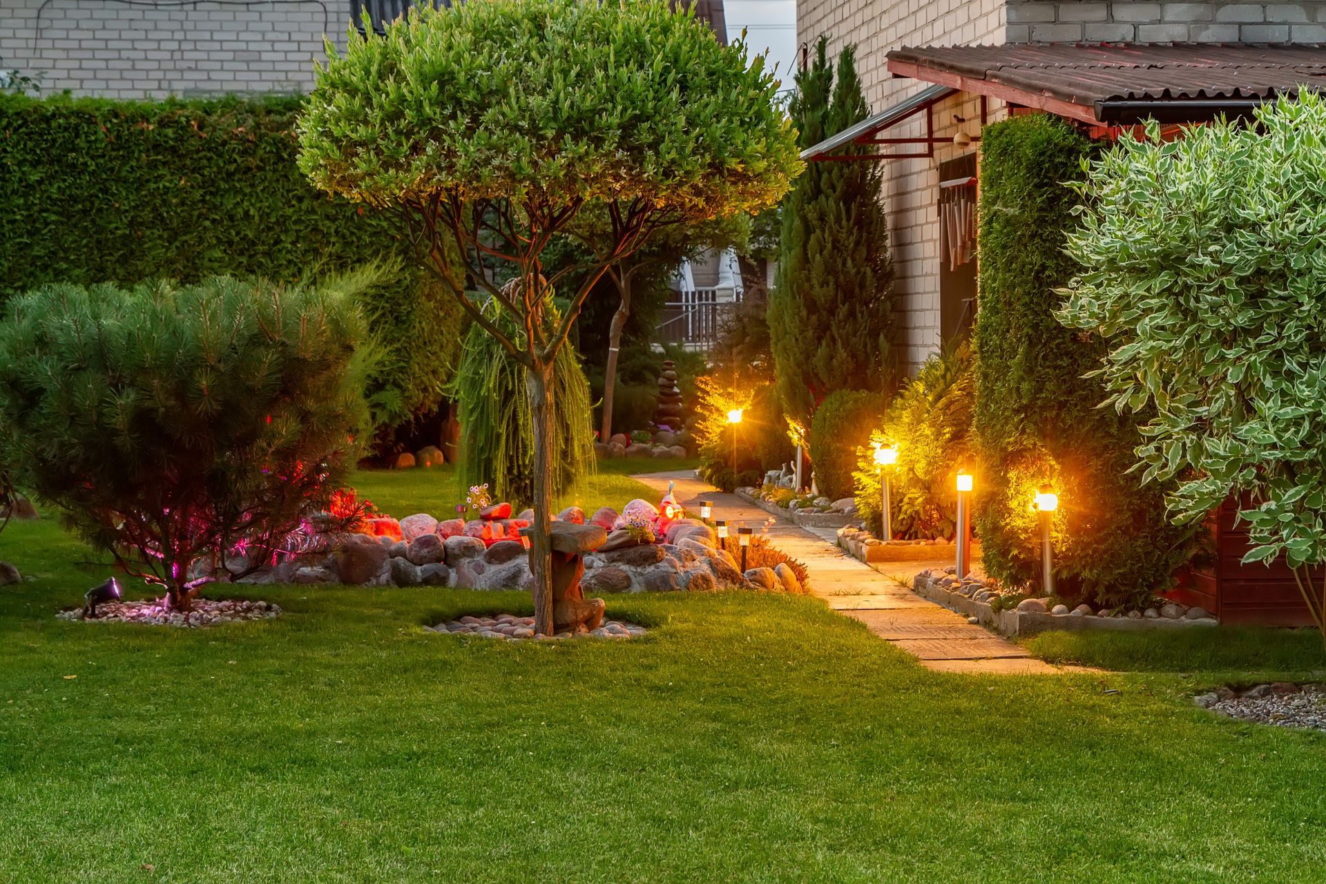 Lush green garden with lighted pathway, trimmed trees, and flower beds. Evening setting with warm lighting.