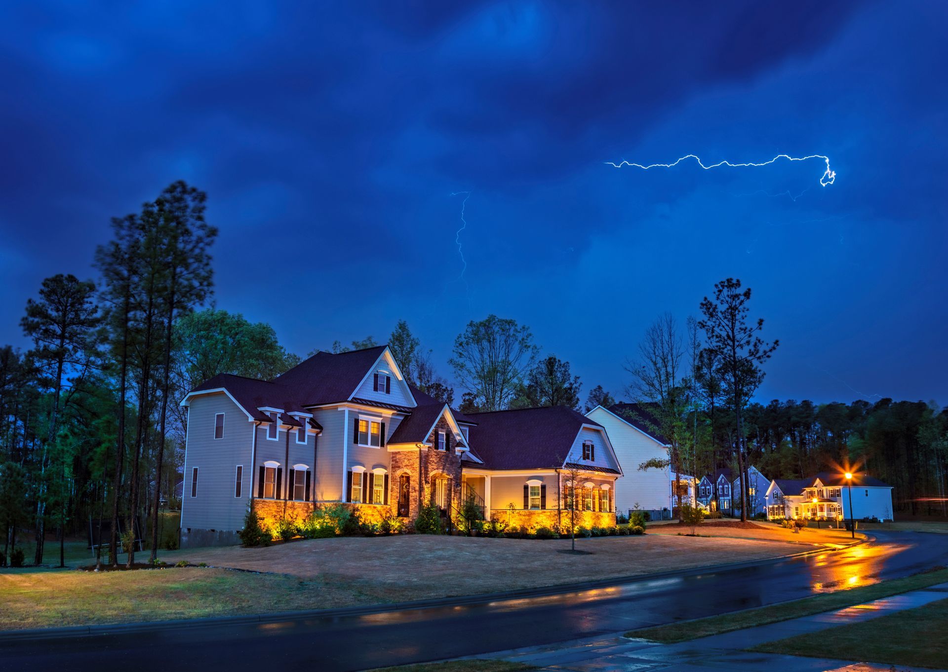 A house illuminated at night during a thunderstorm. Lightning streaks across a dark blue sky.