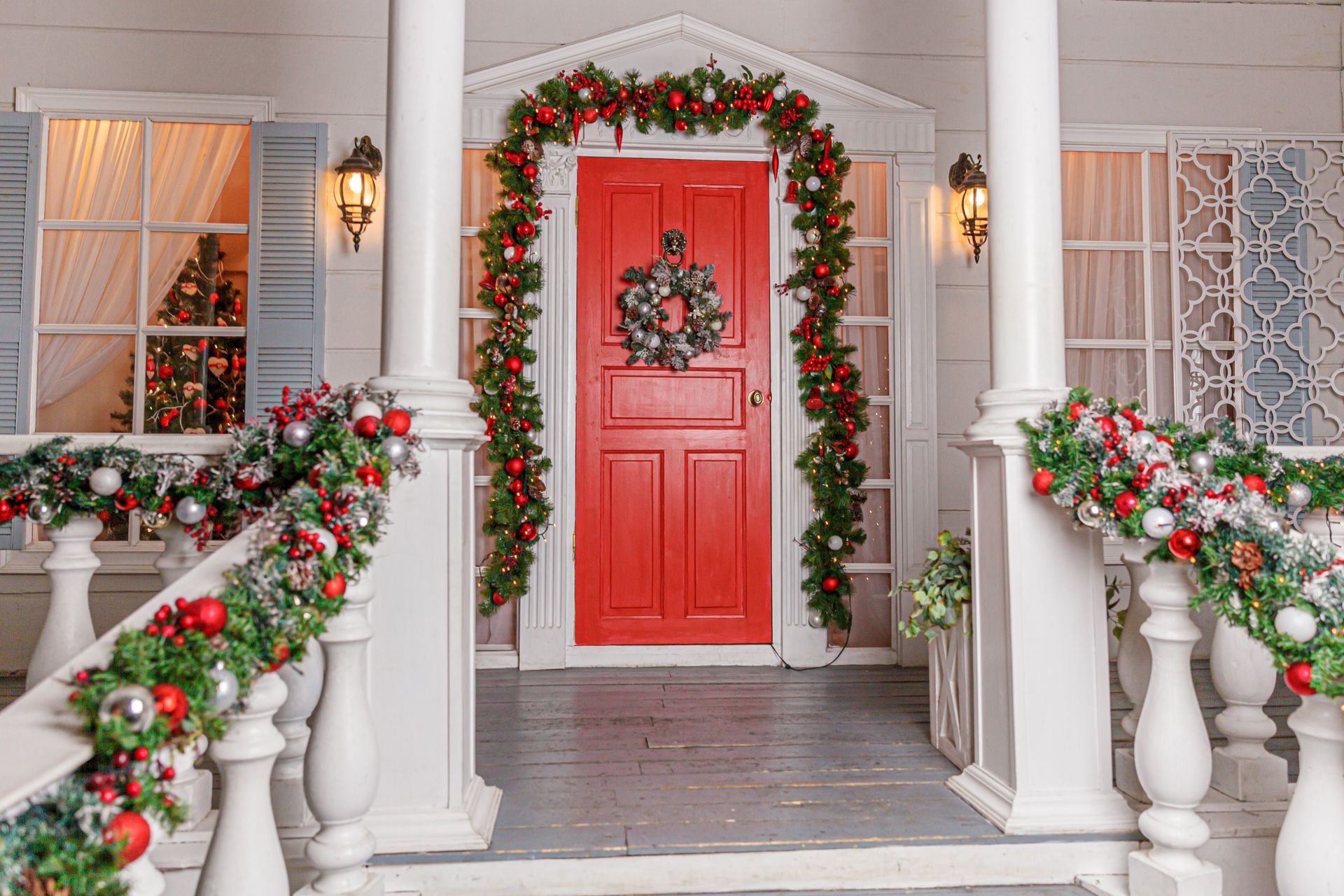 Festive porch with red door, garland, and wreath. White pillars and railing; windows with Christmas trees inside.