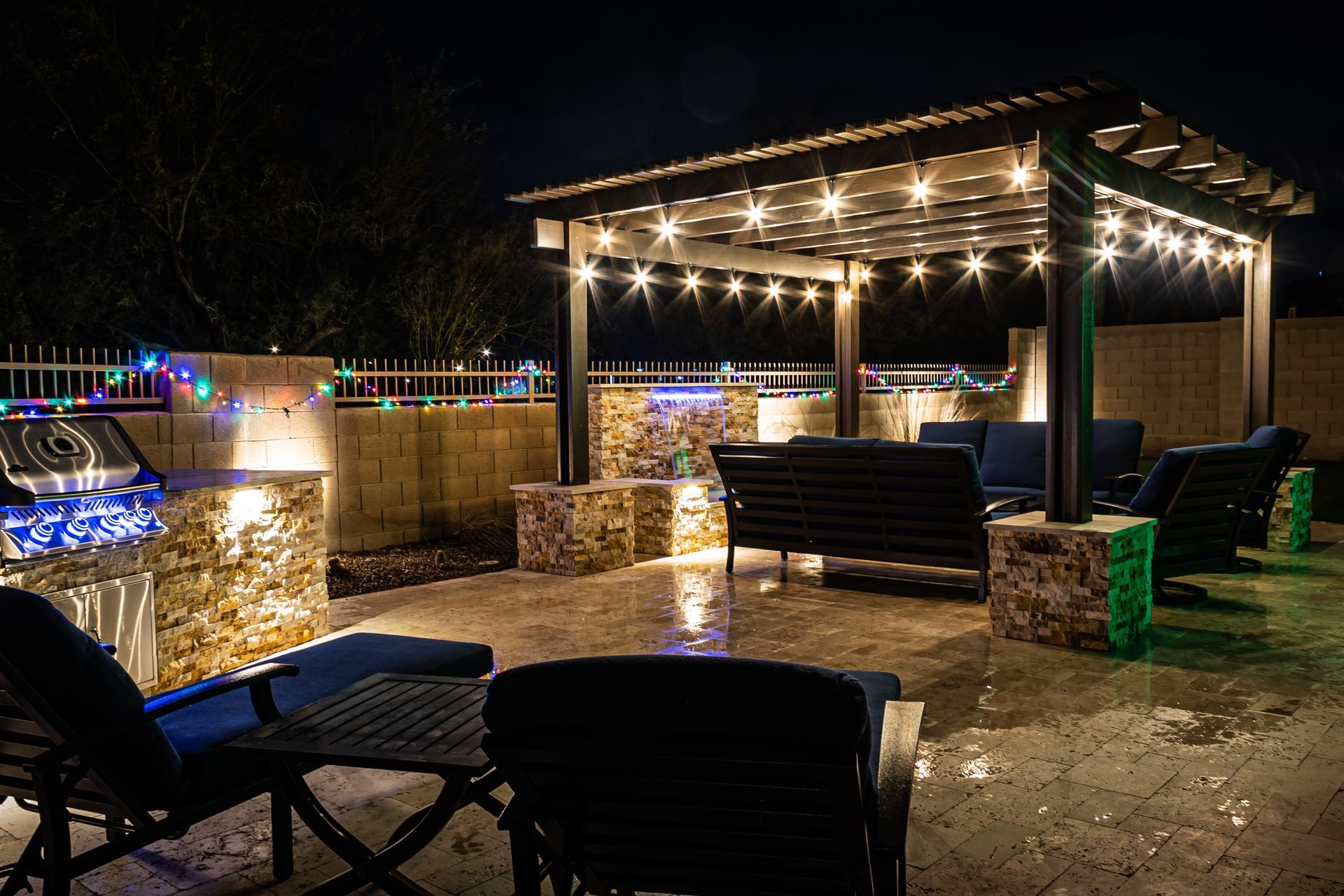 A nighttime patio scene with stone pillars, a lit pergola, comfortable outdoor furniture, and decorative string lights.