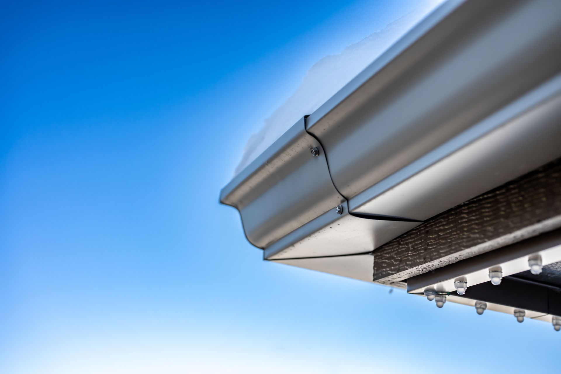A metal gutter corner with a seam is installed along the edge of a roof against a bright blue sky.